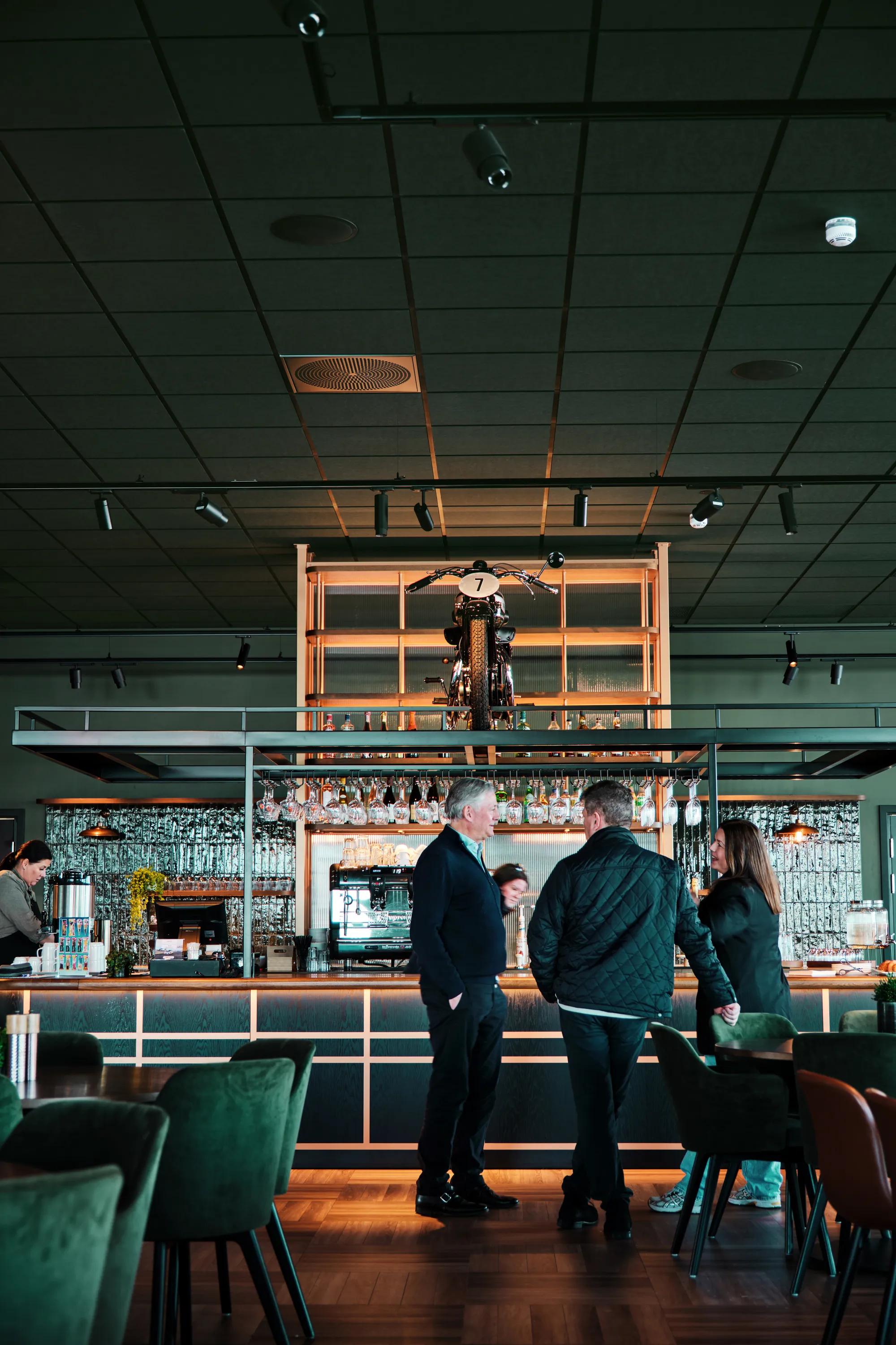 A modern bar interior featuring dark green acoustic ceiling tiles, track lighting, and a display shelf holding a vintage motorcycle above the bar counter. People are gathered at the bar, surrounded by dark green seating and warm wooden finishes.