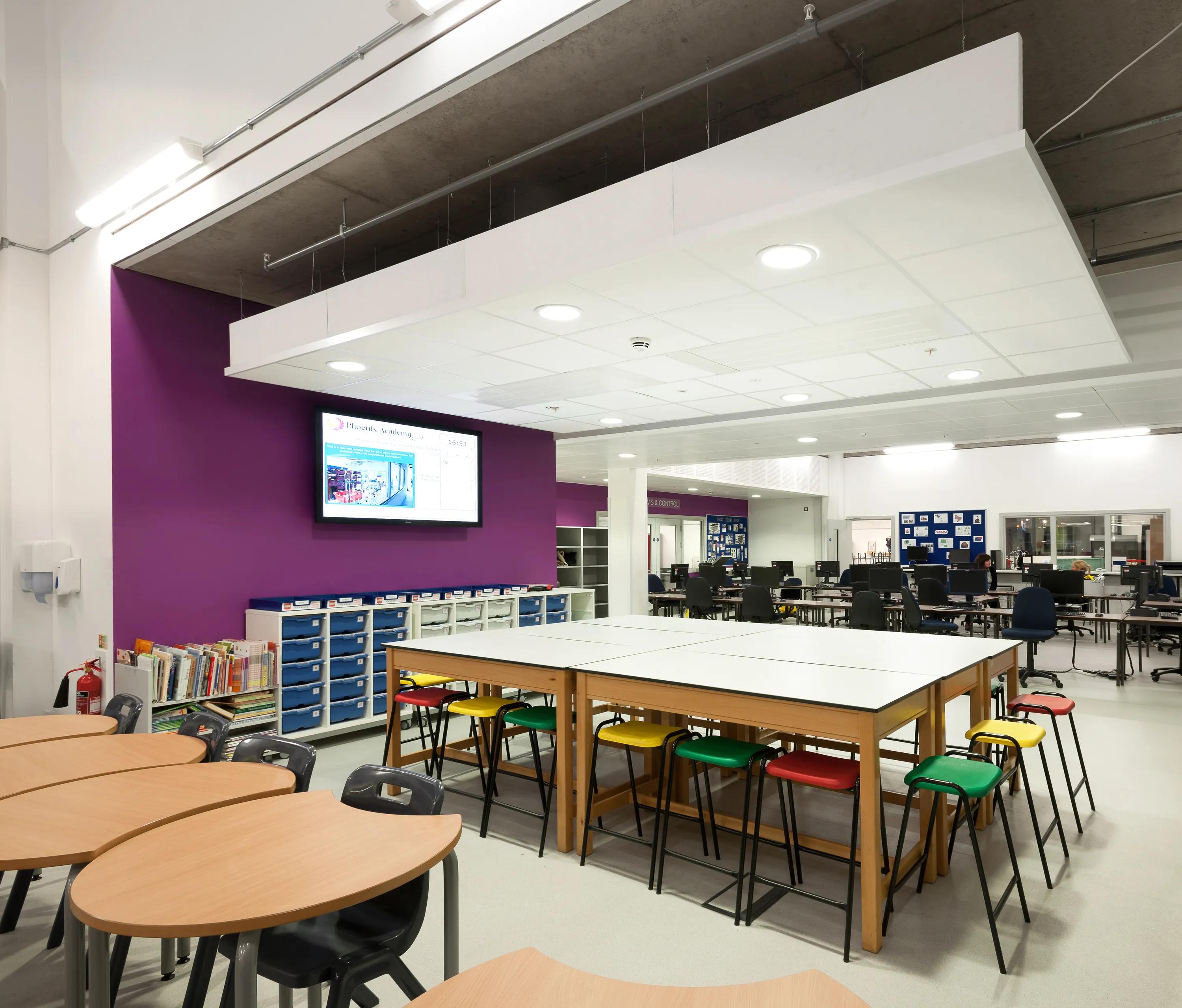 School learning space with group tables, computer workstations, and a suspended white acoustic ceiling island with integrated lighting above the classroom area.