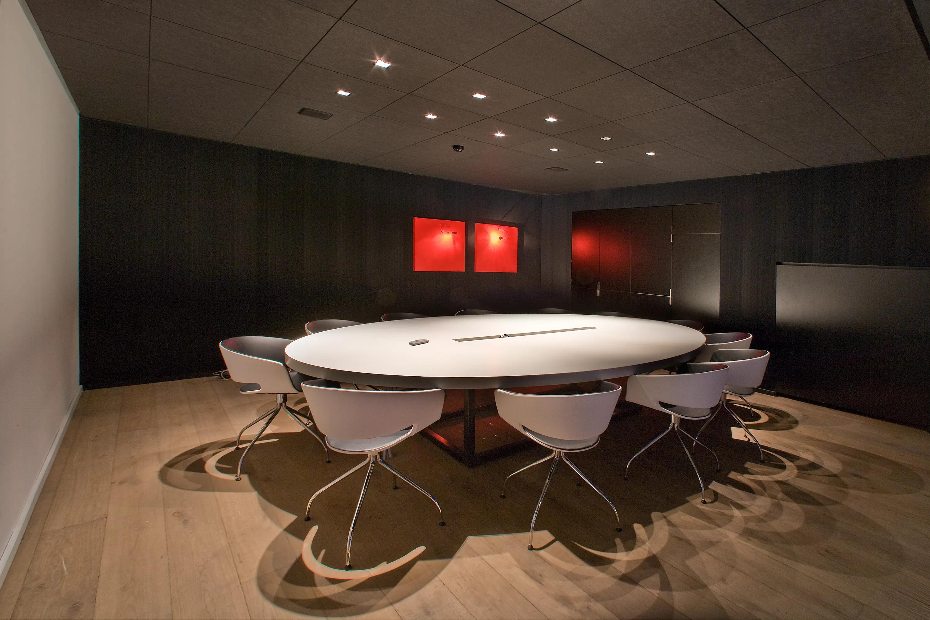 Meeting room with dark acoustic ceiling tiles, round conference table, surrounding chairs, and subdued lighting creating a focused, enclosed workspace.