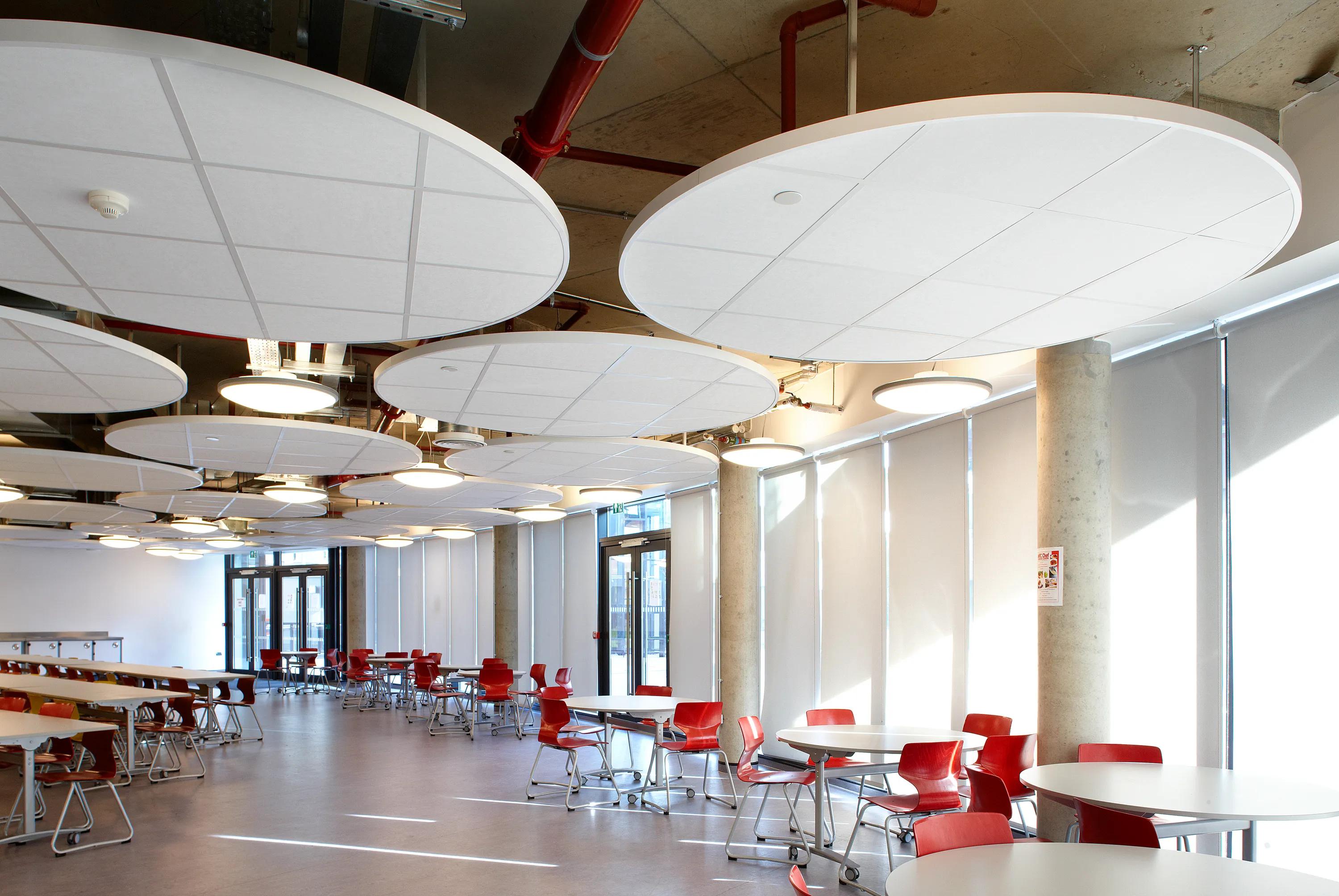 A modern dining area with a floating raft ceiling, round tables, chairs, and large circular ceiling panels. Sunlight streams in through tall windows.
