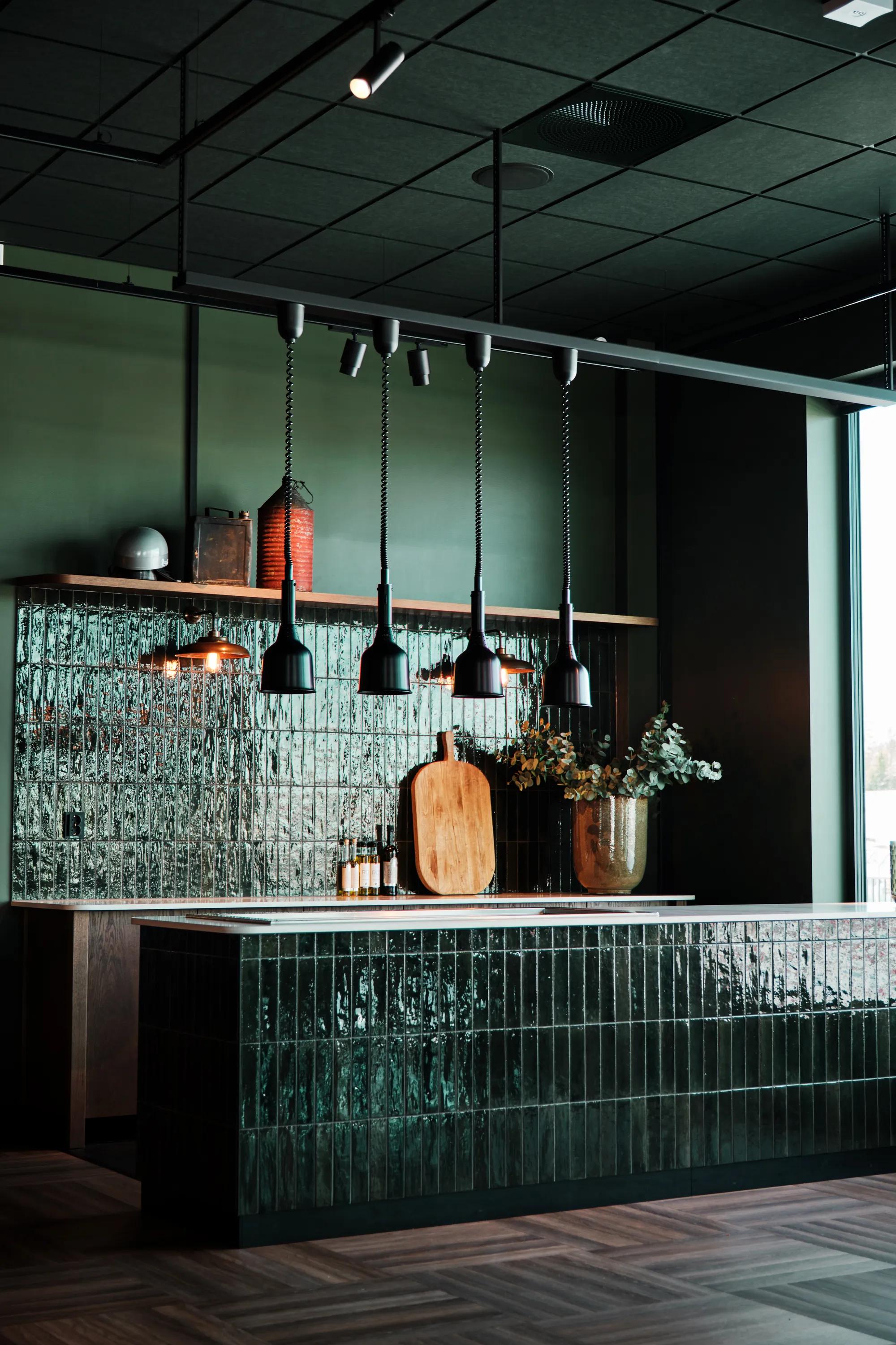 Modern kitchen with dark green tiled island, pendant lights, wooden cutting boards, and a vase with plants, set against textured walls.