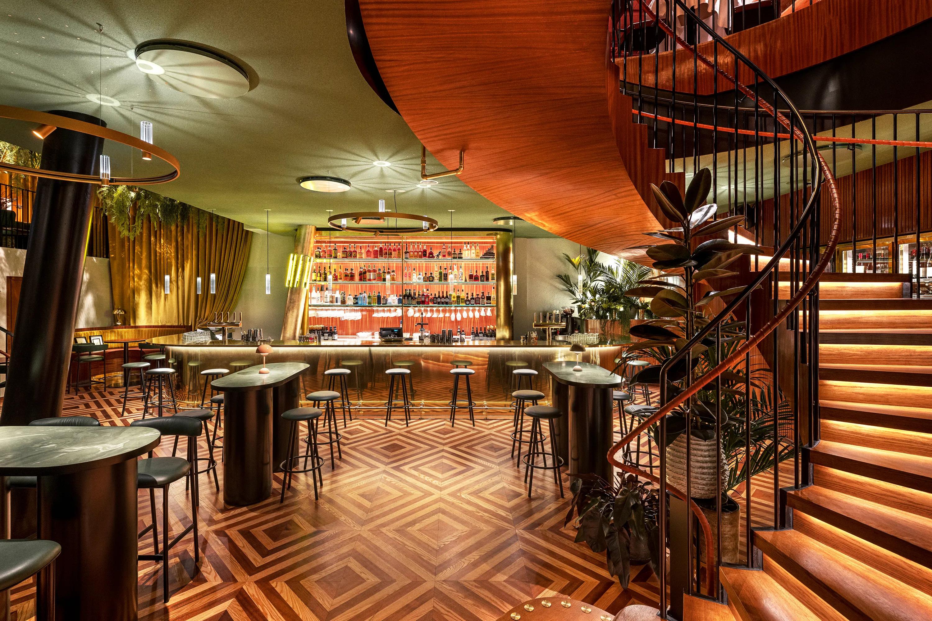 A stylish bar with patterned wooden floors, a spiral staircase, high tables, stools, and a well-stocked, illuminated bar counter in the background.
