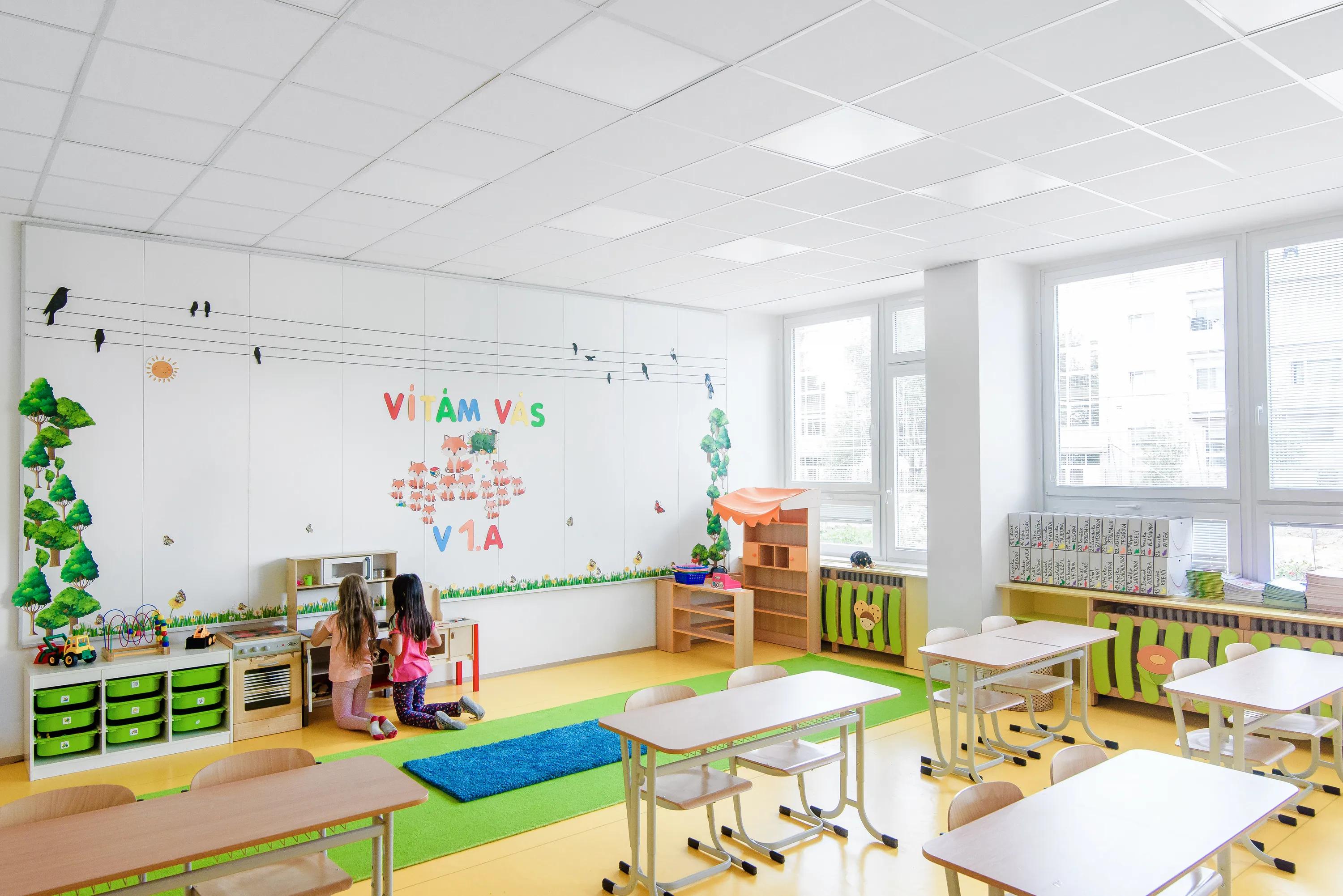 Primary school classroom with white acoustic ceiling tiles, colourful learning areas, desks and chairs, and children playing in a soft seating corner by the wall.