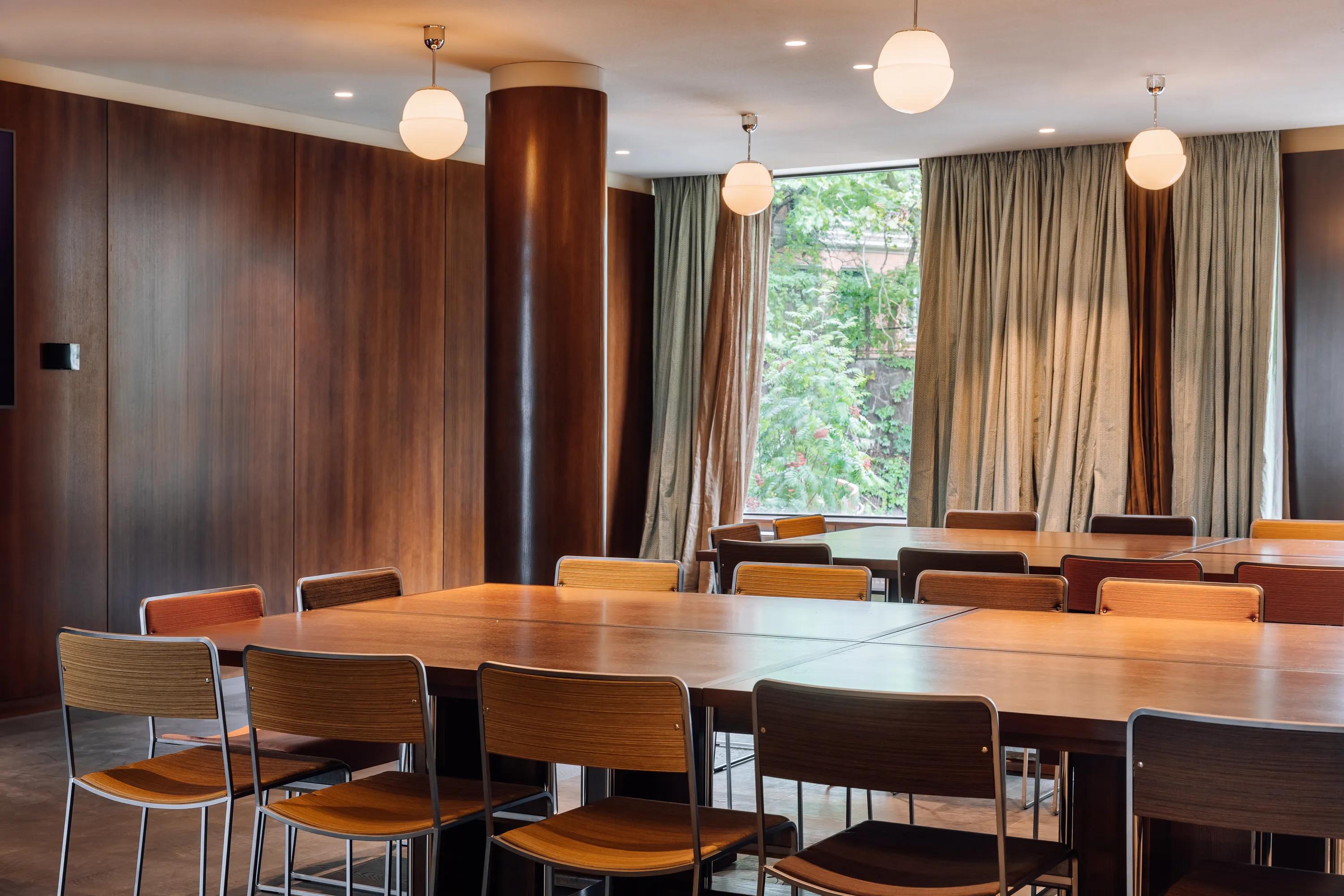 Meeting room with a smooth monolithic acoustic ceiling, wooden tables and chairs, pendant lighting, and large windows with curtains overlooking greenery.