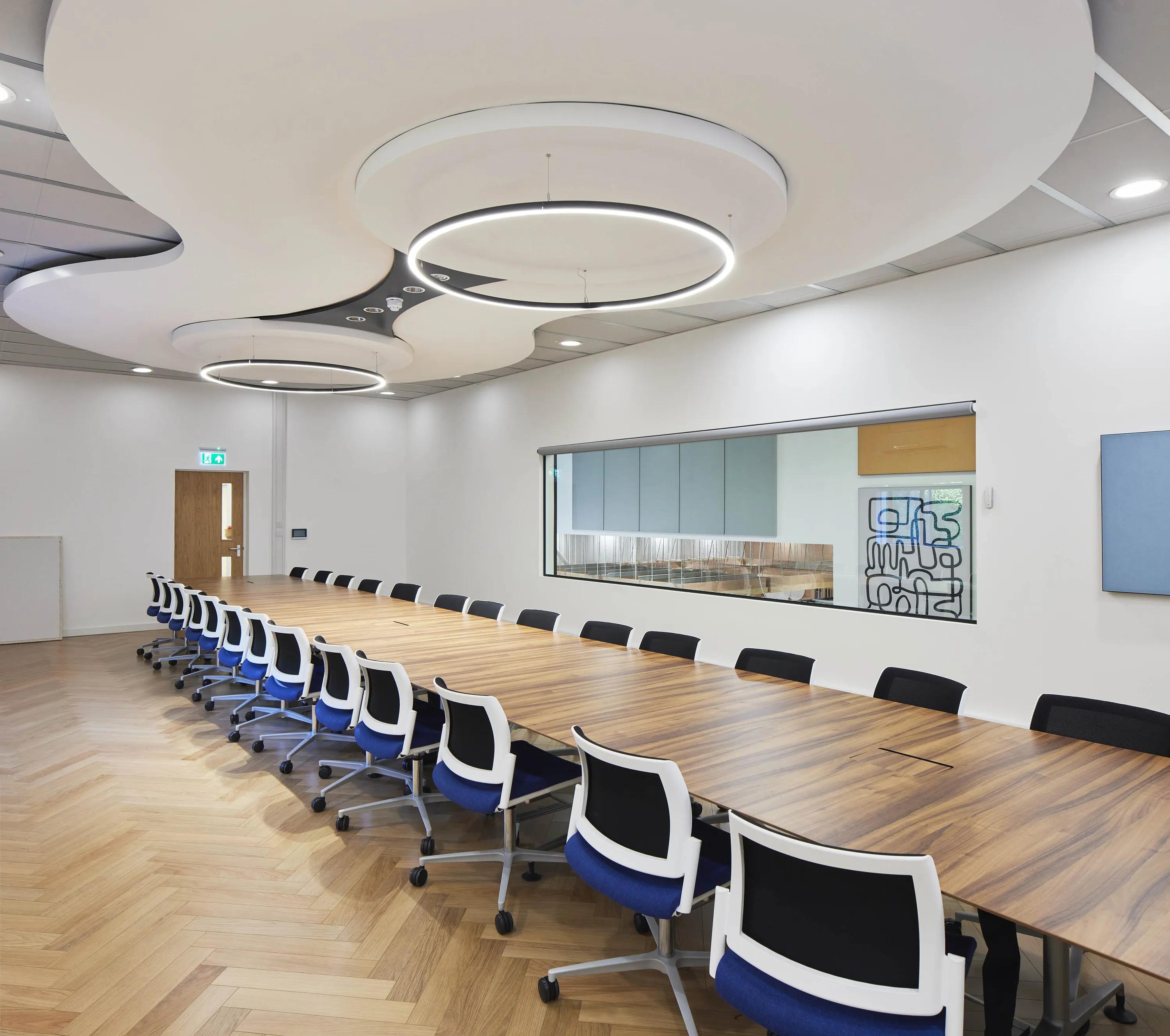 Conference room with a sculpted monolithic acoustic ceiling featuring curved forms and circular lighting, long meeting table, and chairs arranged for group collaboration.