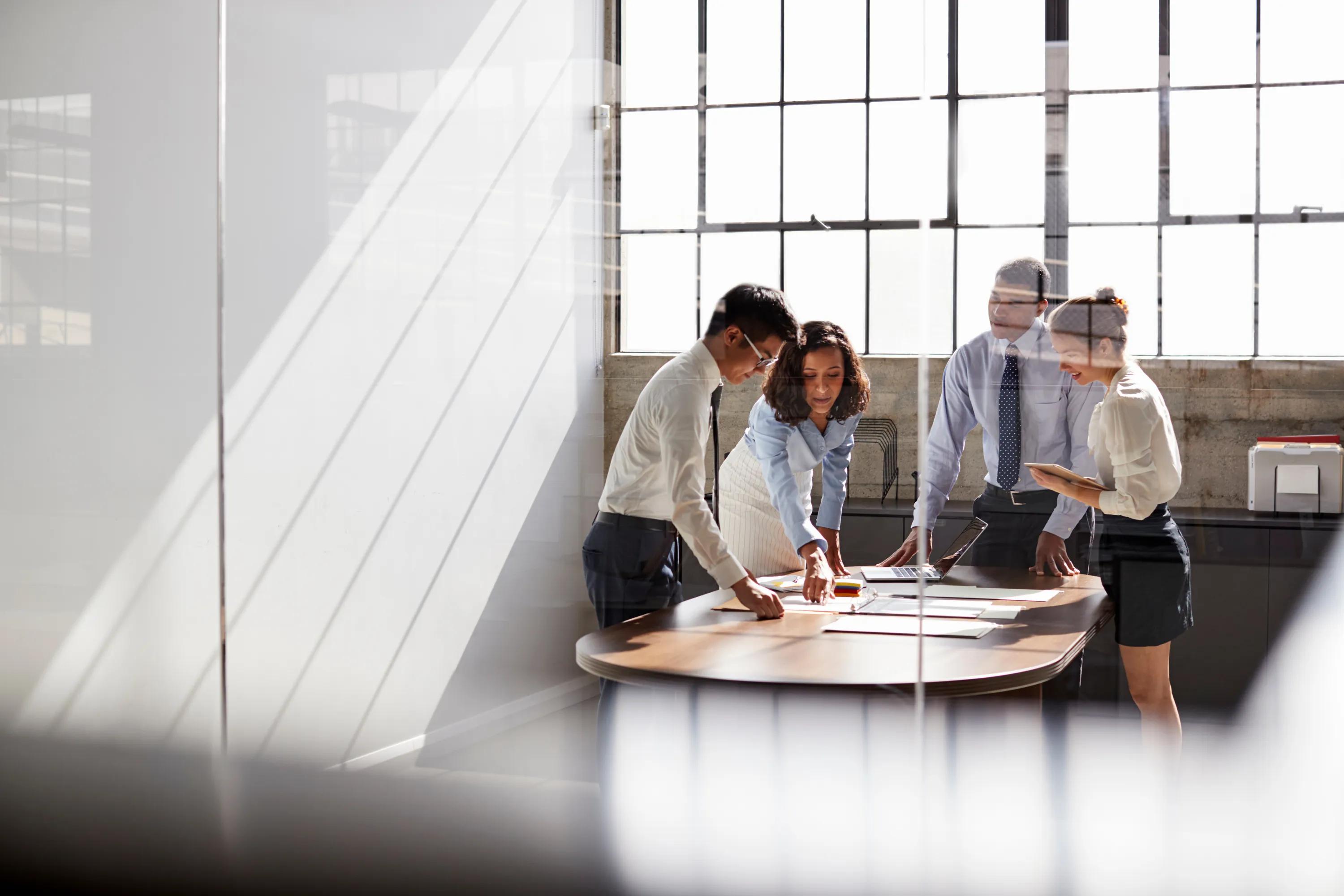 Modern office meeting with four professionals working together at a table in a loft-style room with tall steel-framed windows
