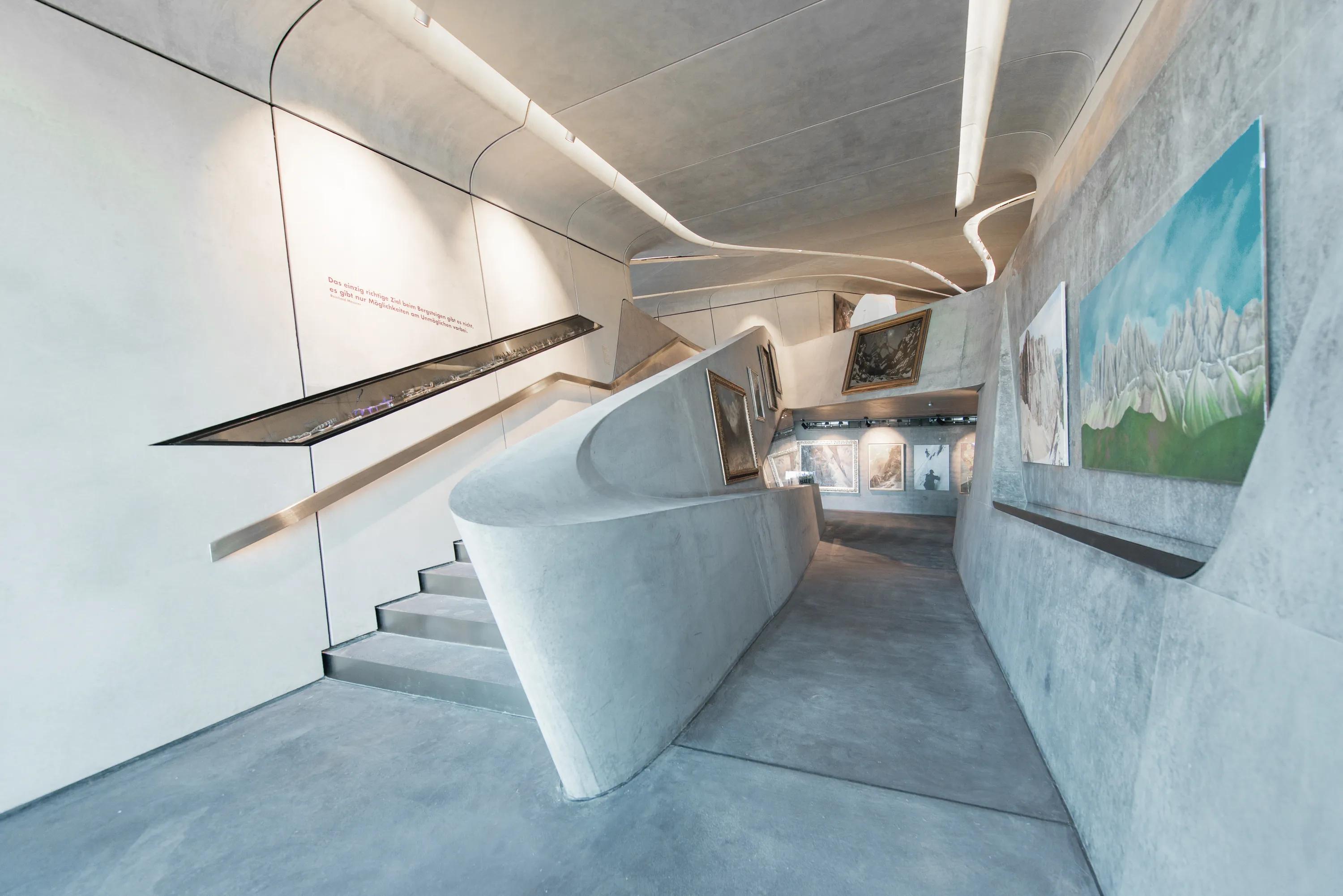 Concrete exhibition space with a large panoramic window overlooking a snowy mountain landscape, displaying sculptural pieces and interpretation panels inside a curved architectural interior.