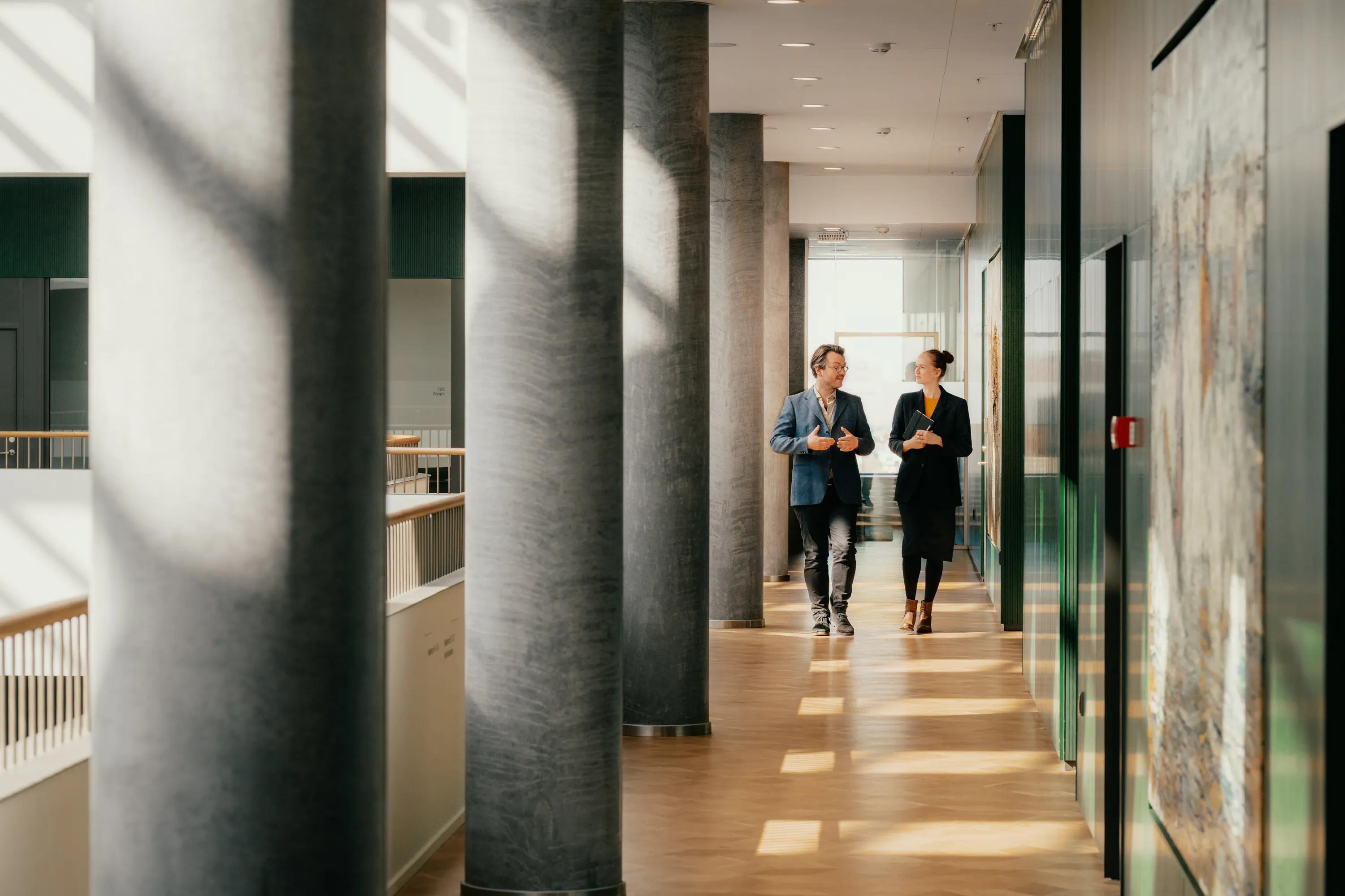 Two people walking and talking in a bright hallway with white acoustic modular ceilings, large columns and artwork, illuminated by sunlight streaming through windows.