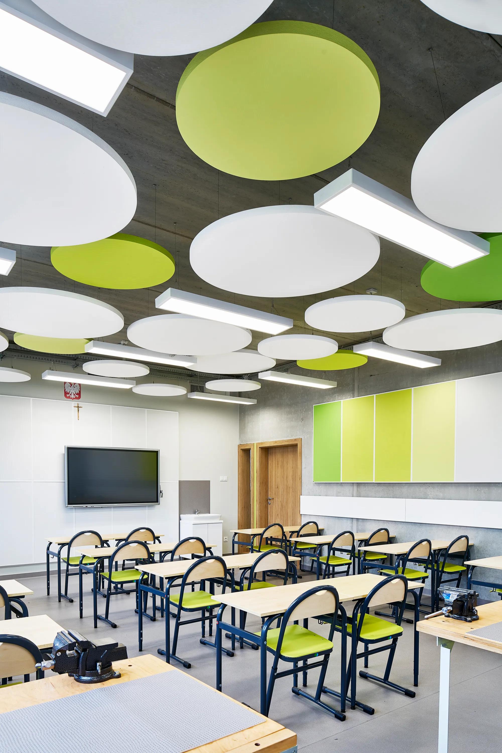 A bright classroom featuring suspended circular acoustic islands in white and lime green, complementing the exposed concrete ceiling and coordinated wall panels.