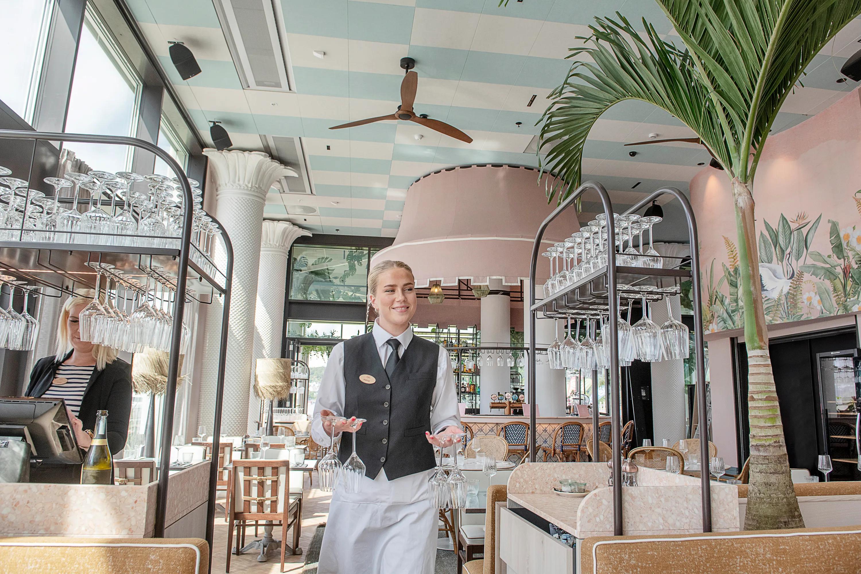 Restaurant interior with a patterned acoustic ceiling and ceiling fans above a bright dining area, where a server carries wine glasses between tables and bar carts.