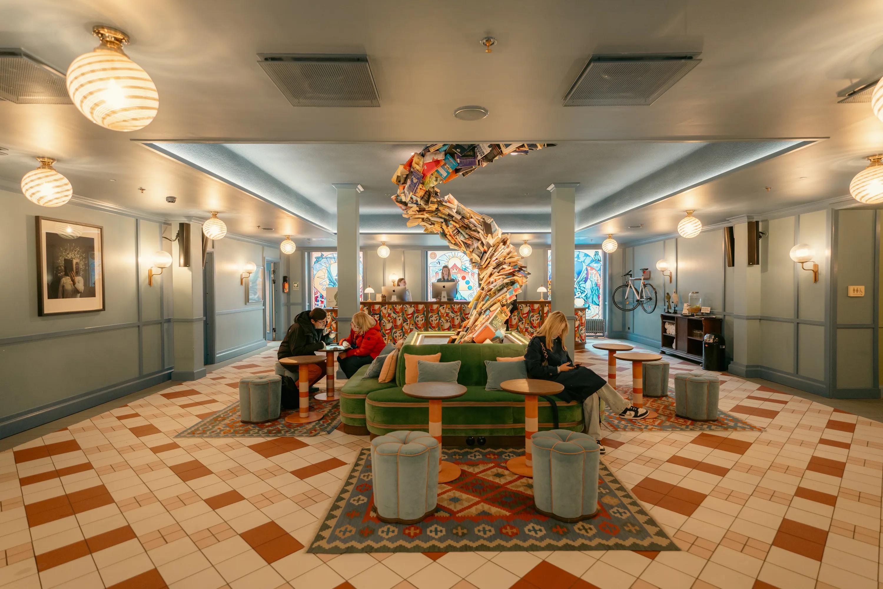 Hotel lobby lounge with a smooth monolithic acoustic ceiling, patterned tile floor, green seating, and a sculptural installation made from stacked books at the centre of the space.