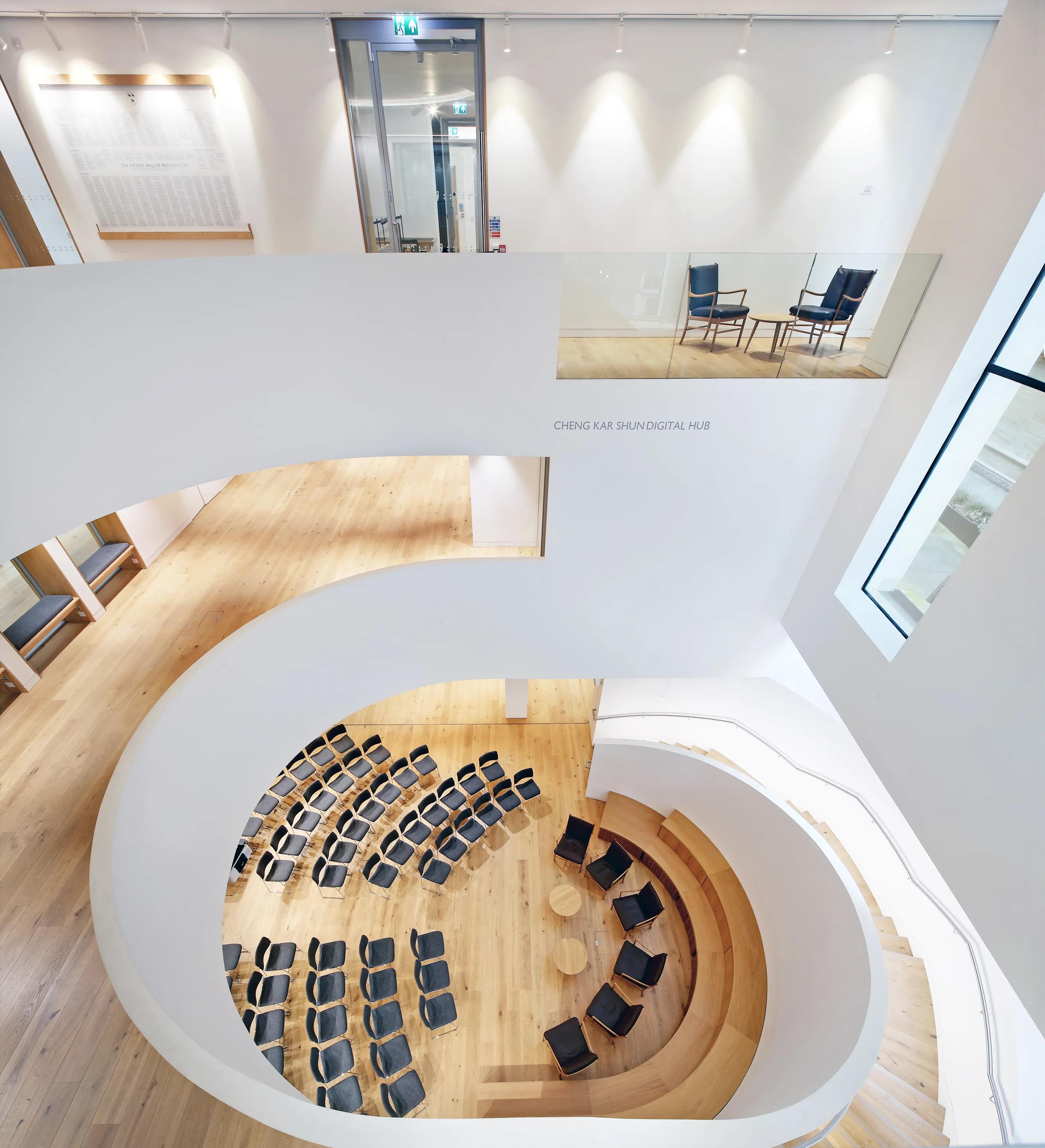 A light-filled atrium featuring curved white acoustic ceilings, a circular tiered seating area with wooden steps, and rows of black chairs arranged for presentations.