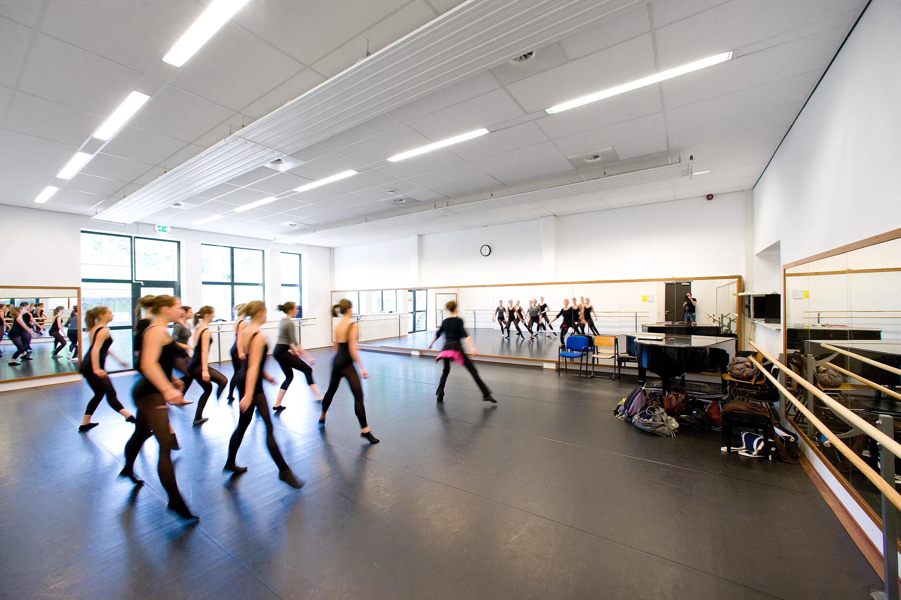 Dance studio with white acoustic ceiling tiles and suspended baffles, mirrored wall, and a group of dancers rehearsing across the open floor.