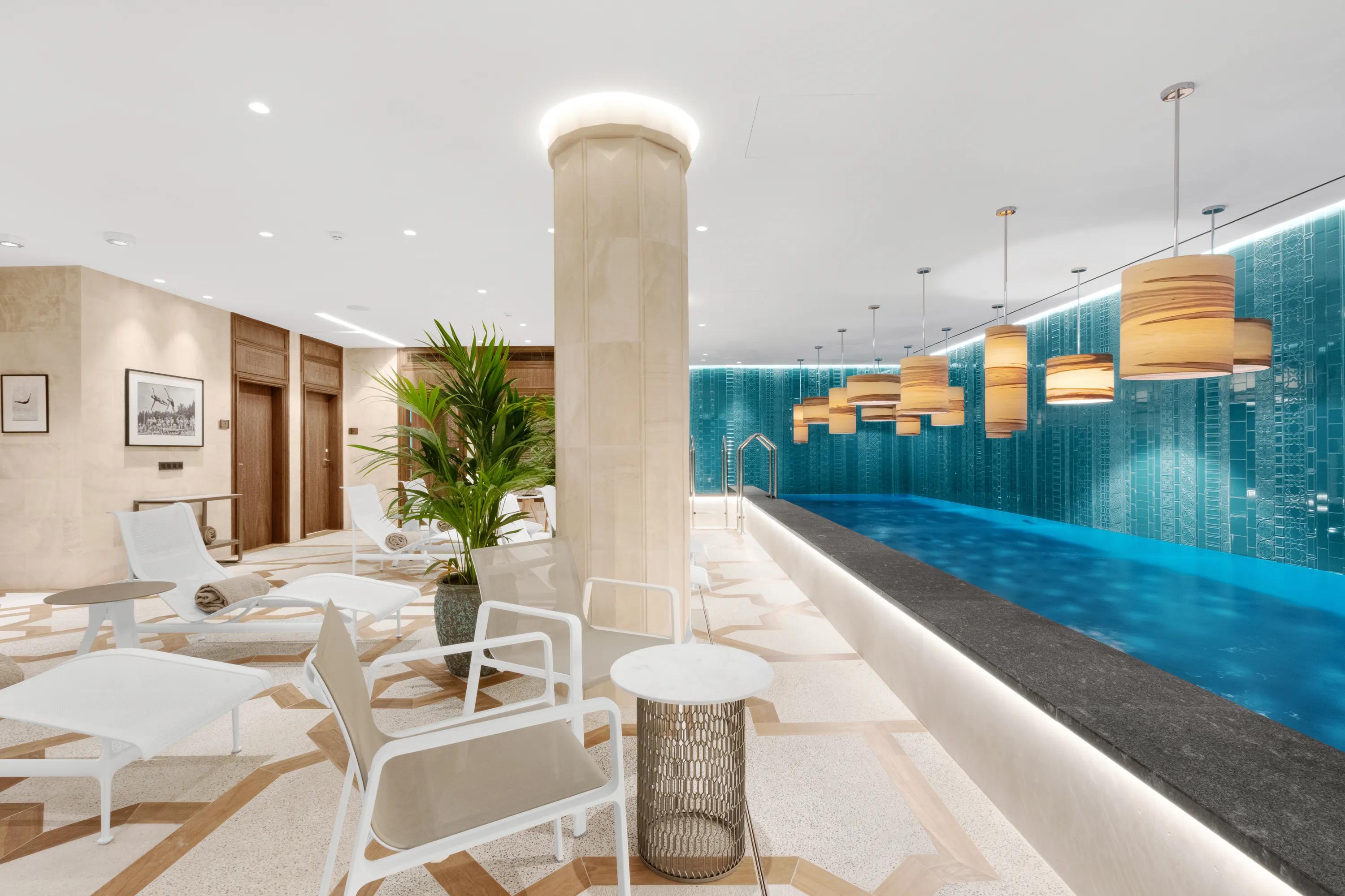 Indoor spa pool area with lounge chairs, turquoise tiled walls, pendant lighting, and a smooth white acoustic ceiling with recessed lights above relaxation seating.