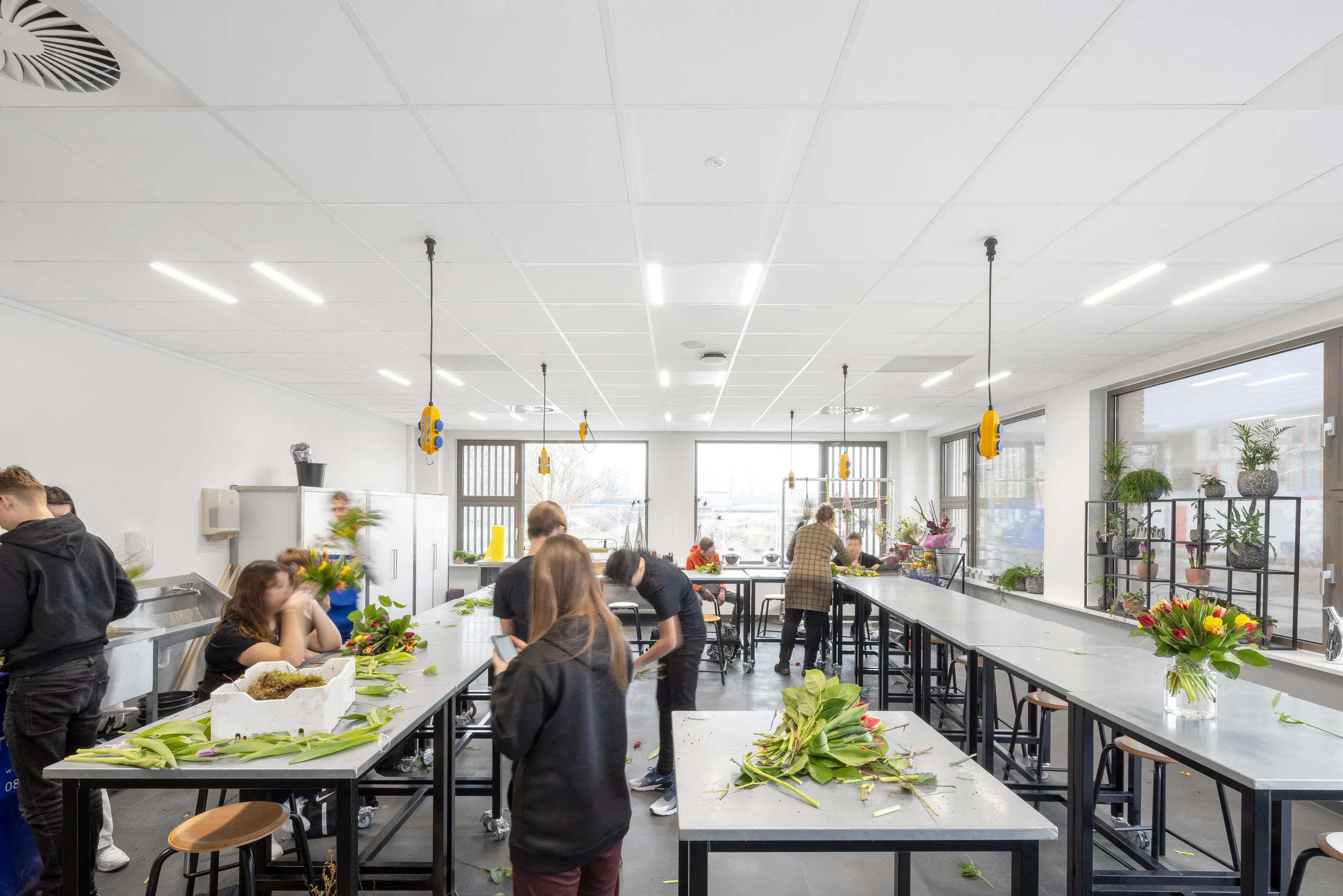 People arranging flowers in a bright, spacious room with white tabletops and large windows. Plants and bouquets adorn the space.
