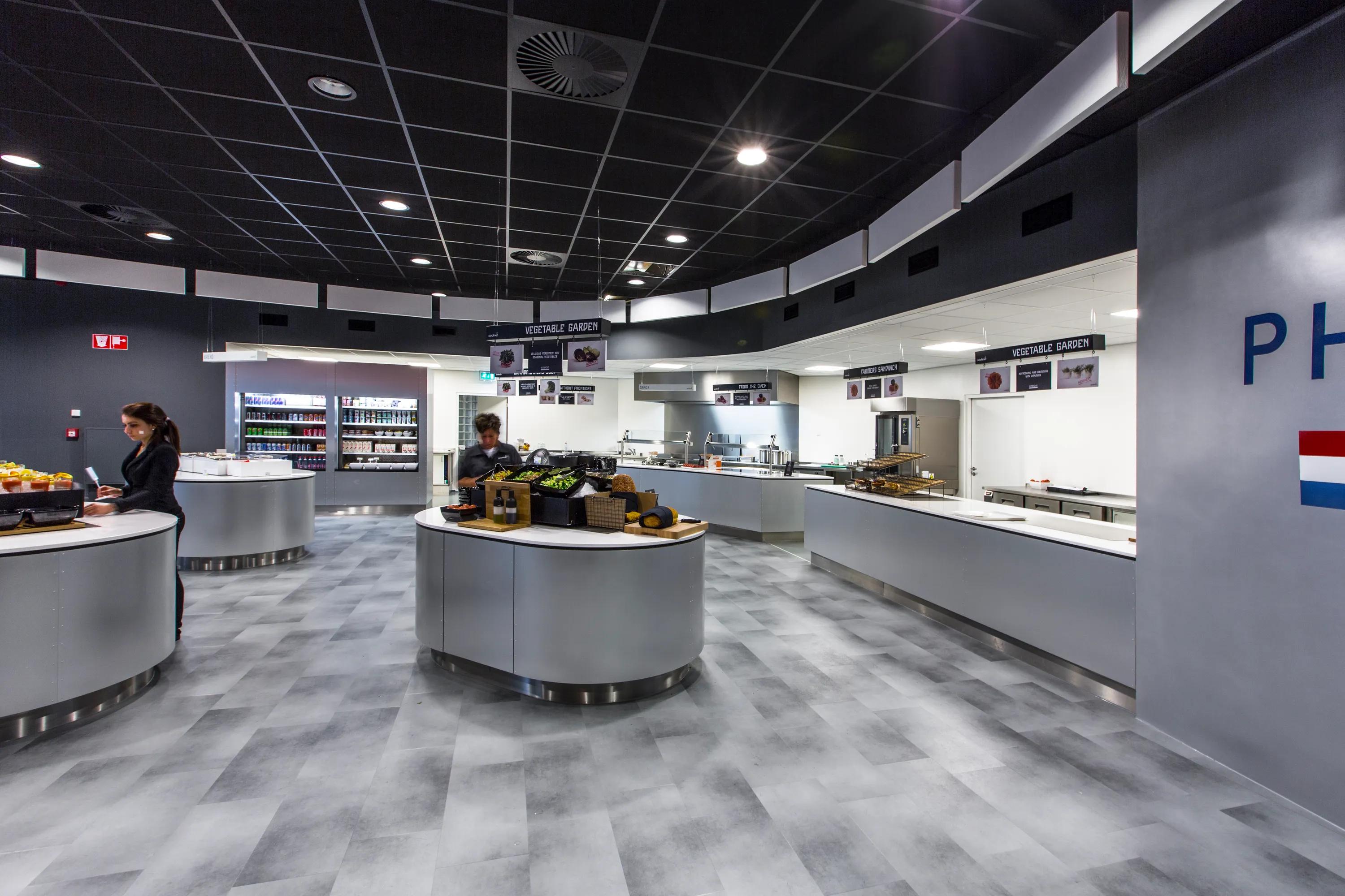 Self-service restaurant interior with food counters, circular serving stations, and a black acoustic grid ceiling above the dining area.