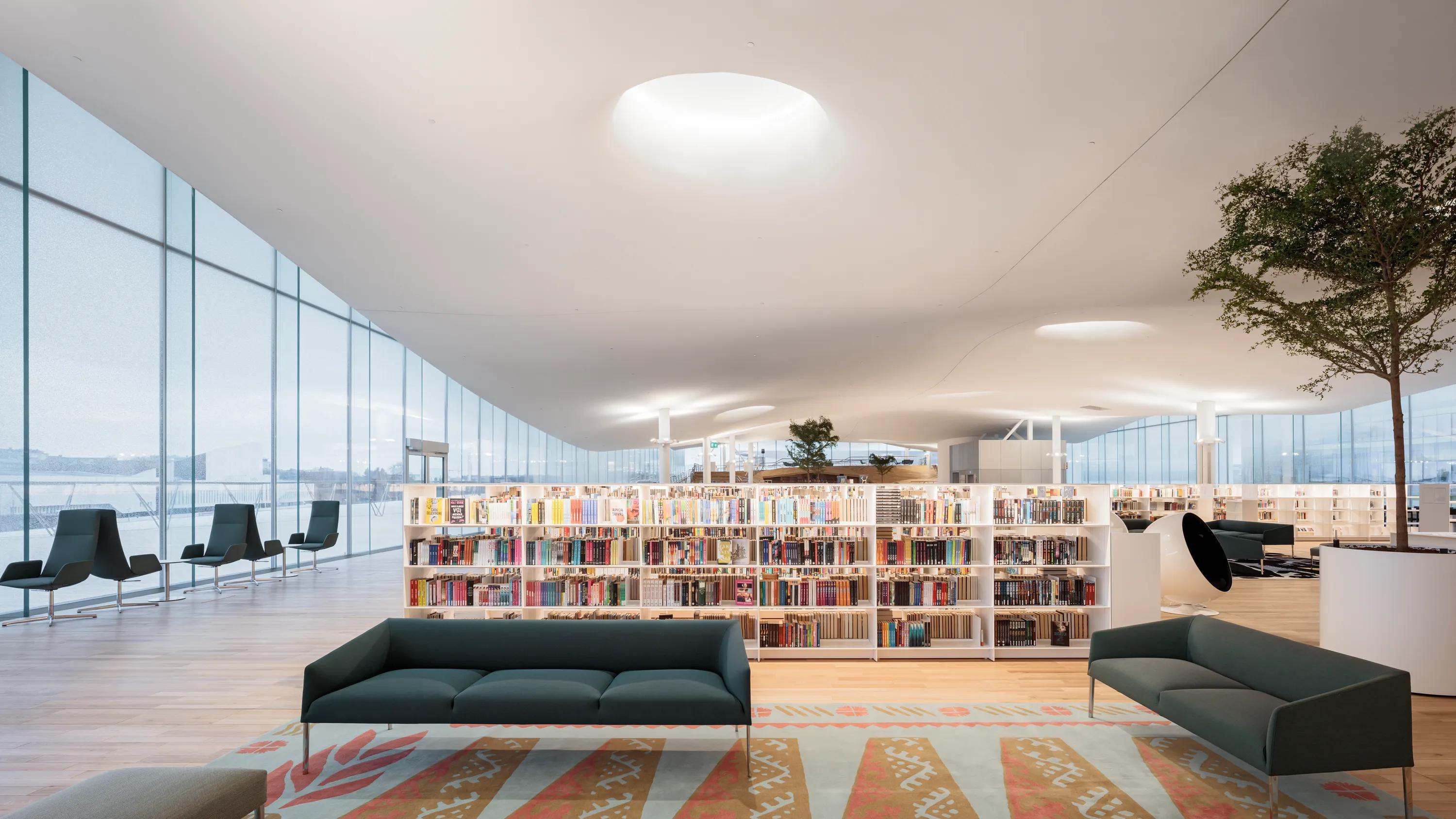 A bright public library with a seamless white acoustic ceiling, large glazed façade, and wide timber steps filled with book.