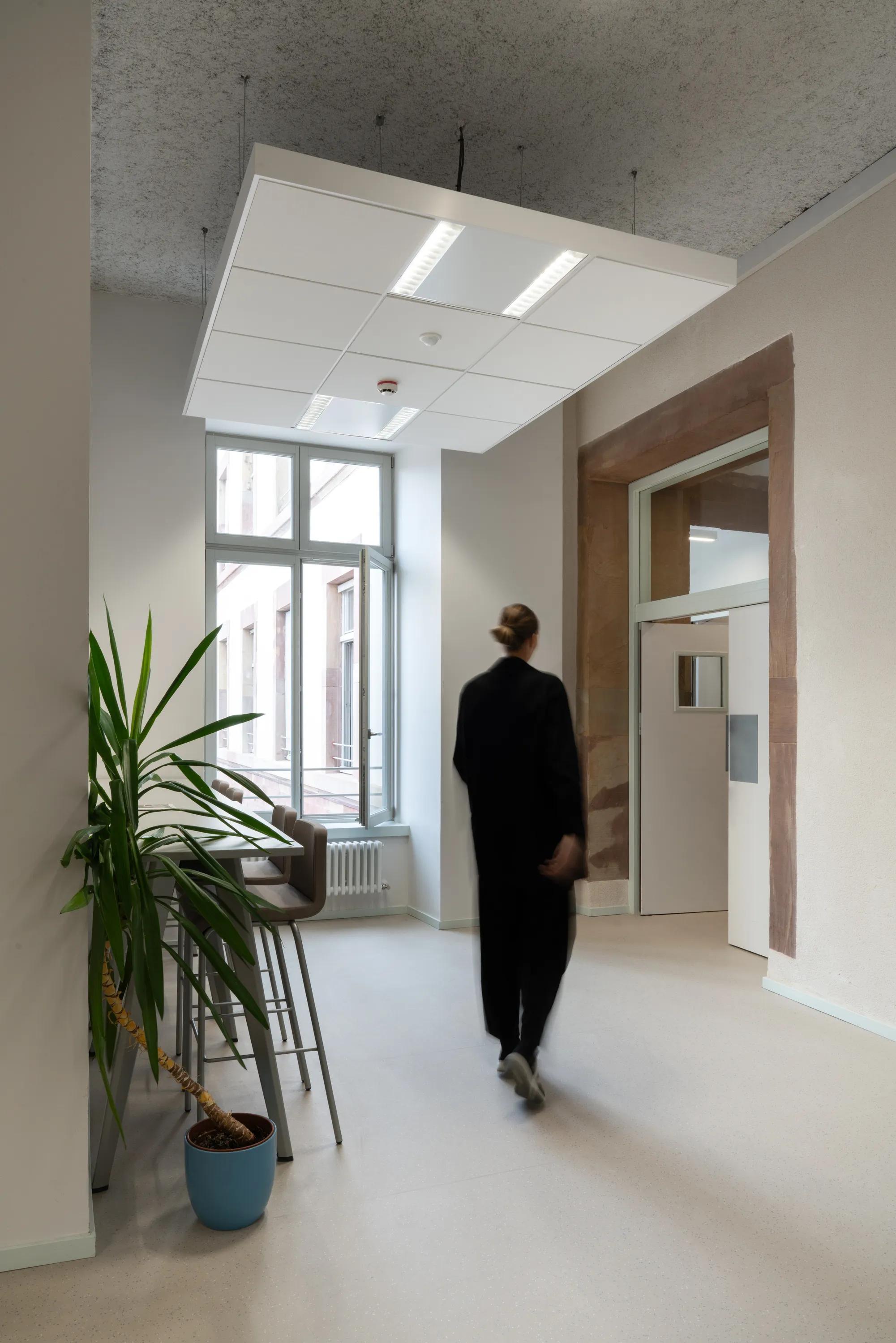 Person in black outfit walking in a modern, minimalist office hallway with a large window and floating raft ceiling