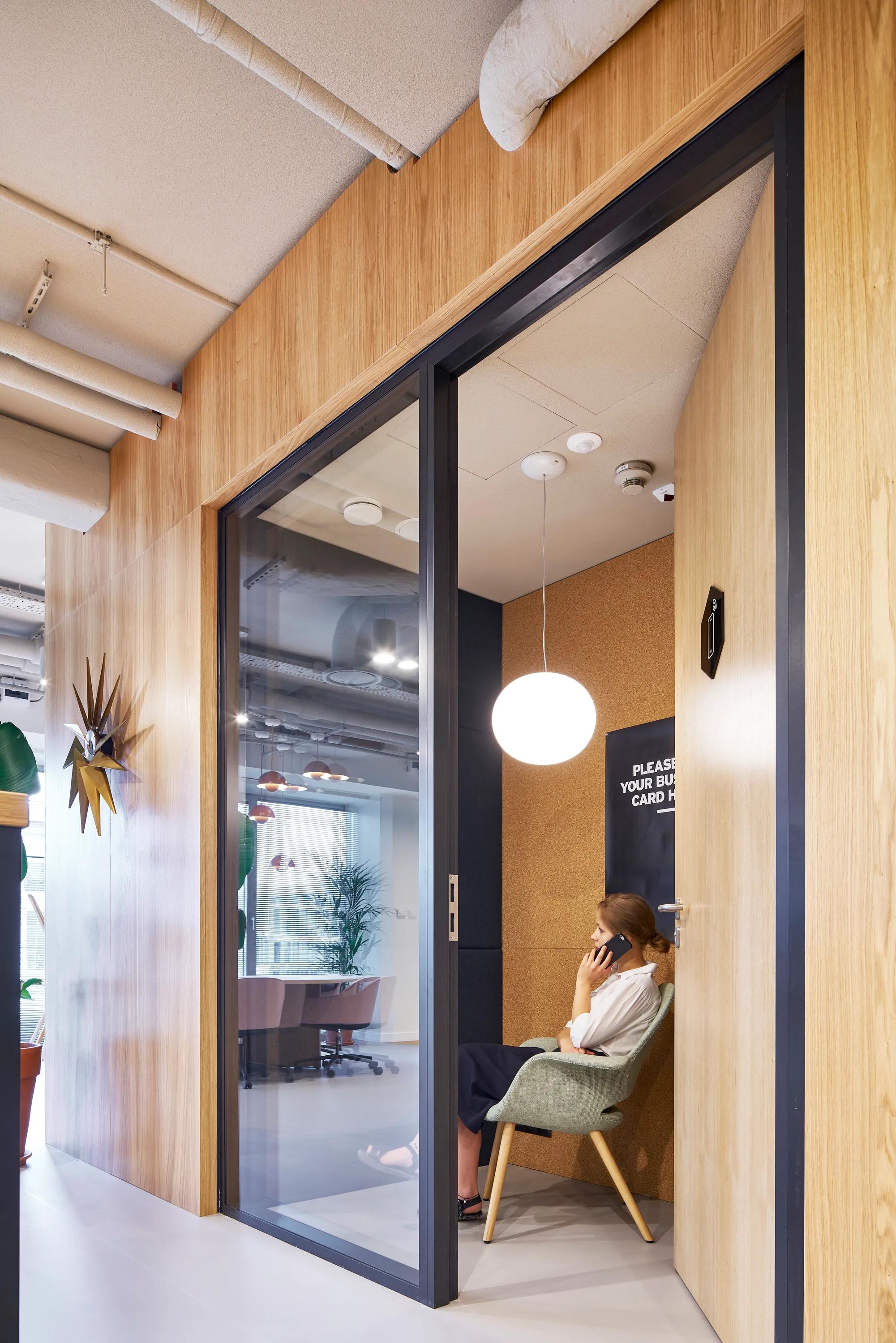 Phone booth area with acoustic ceiling panels and wooden walls, where a person sits in a lounge chair making a call