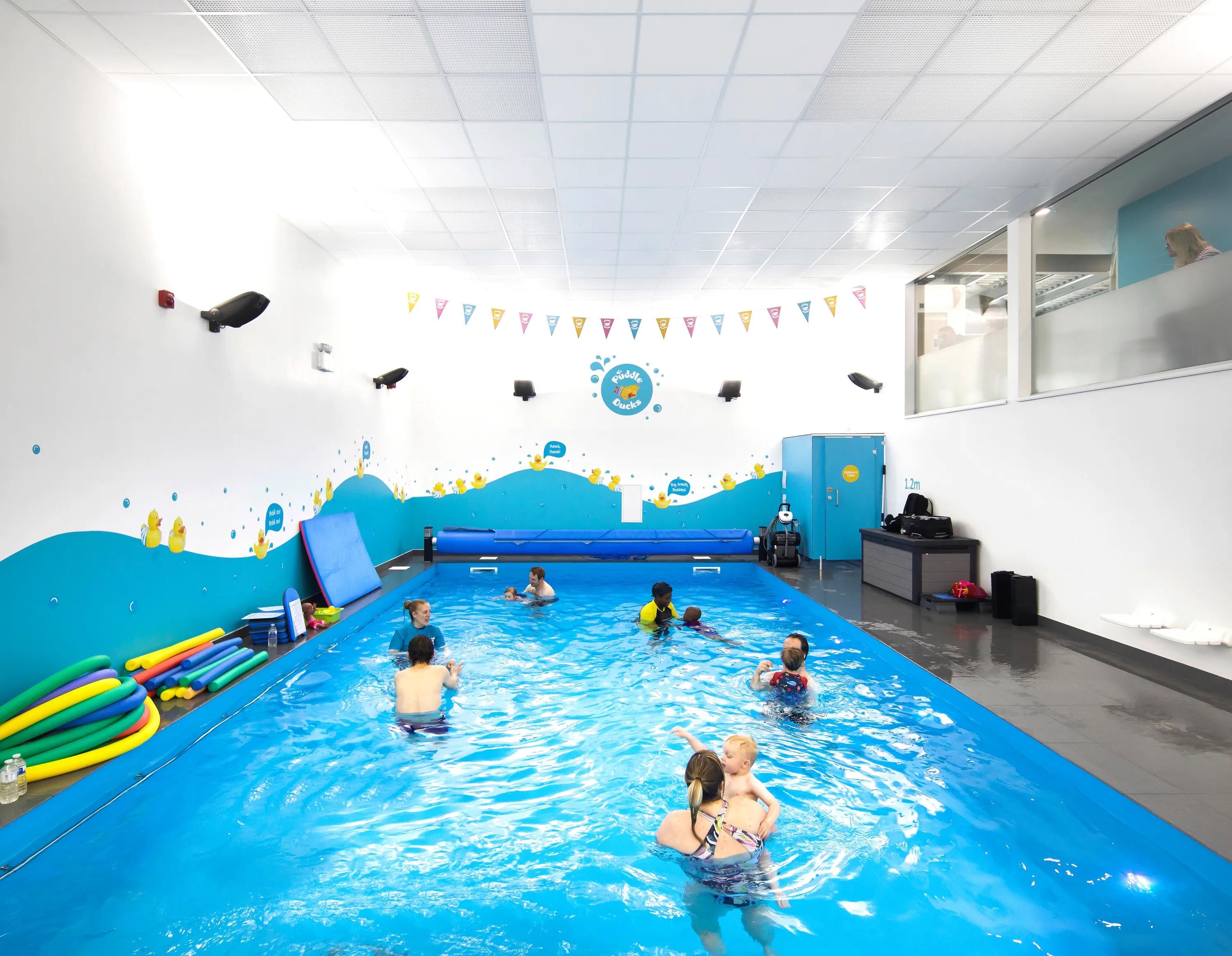 Indoor swimming pool with children and instructor, colorful wall decorations, and pool toys around. Bright overhead lighting and glass observation area.