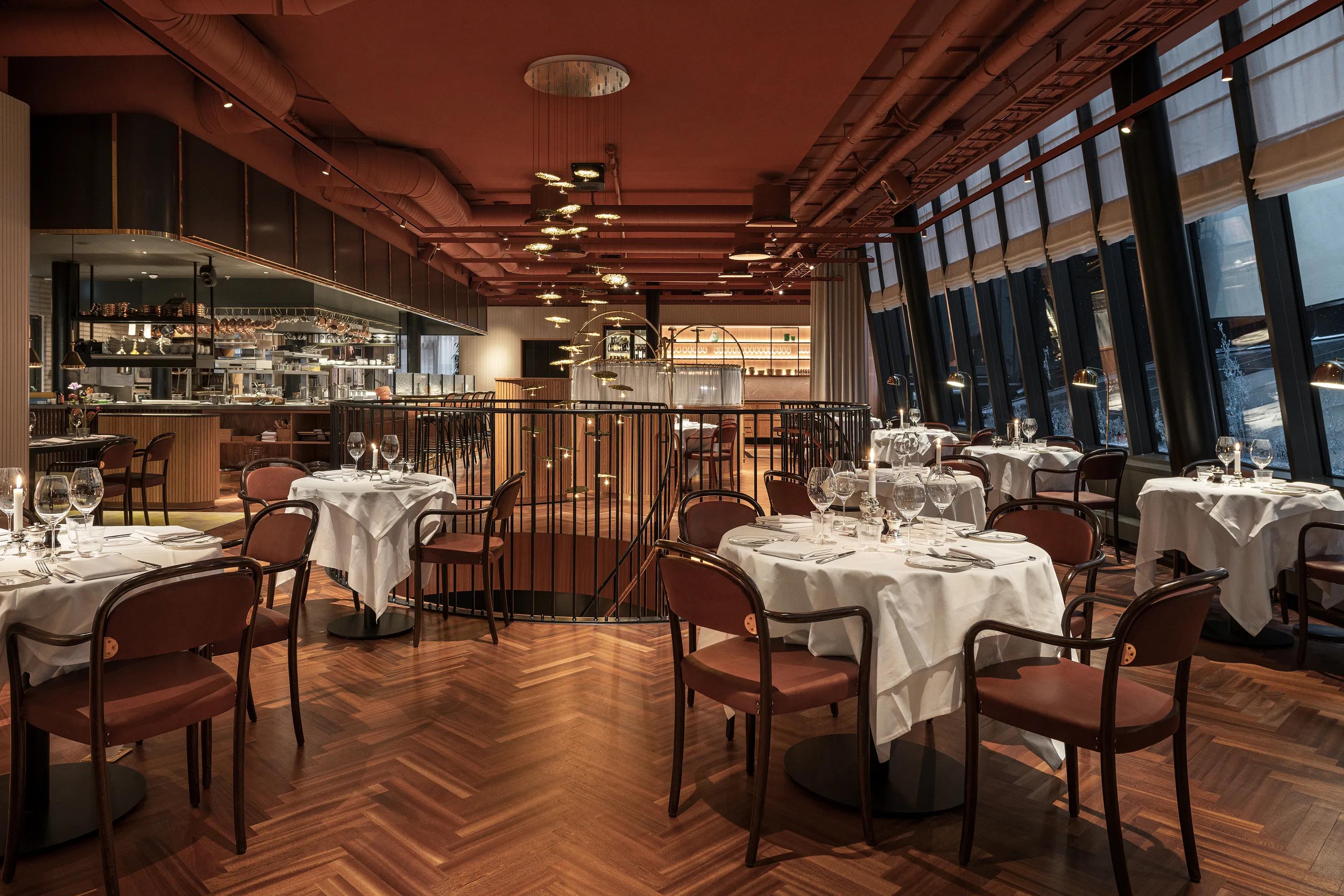 Restaurant interior with a dark maroon monolithic acoustic ceiling, set dining tables, open kitchen, and large windows lining the space.