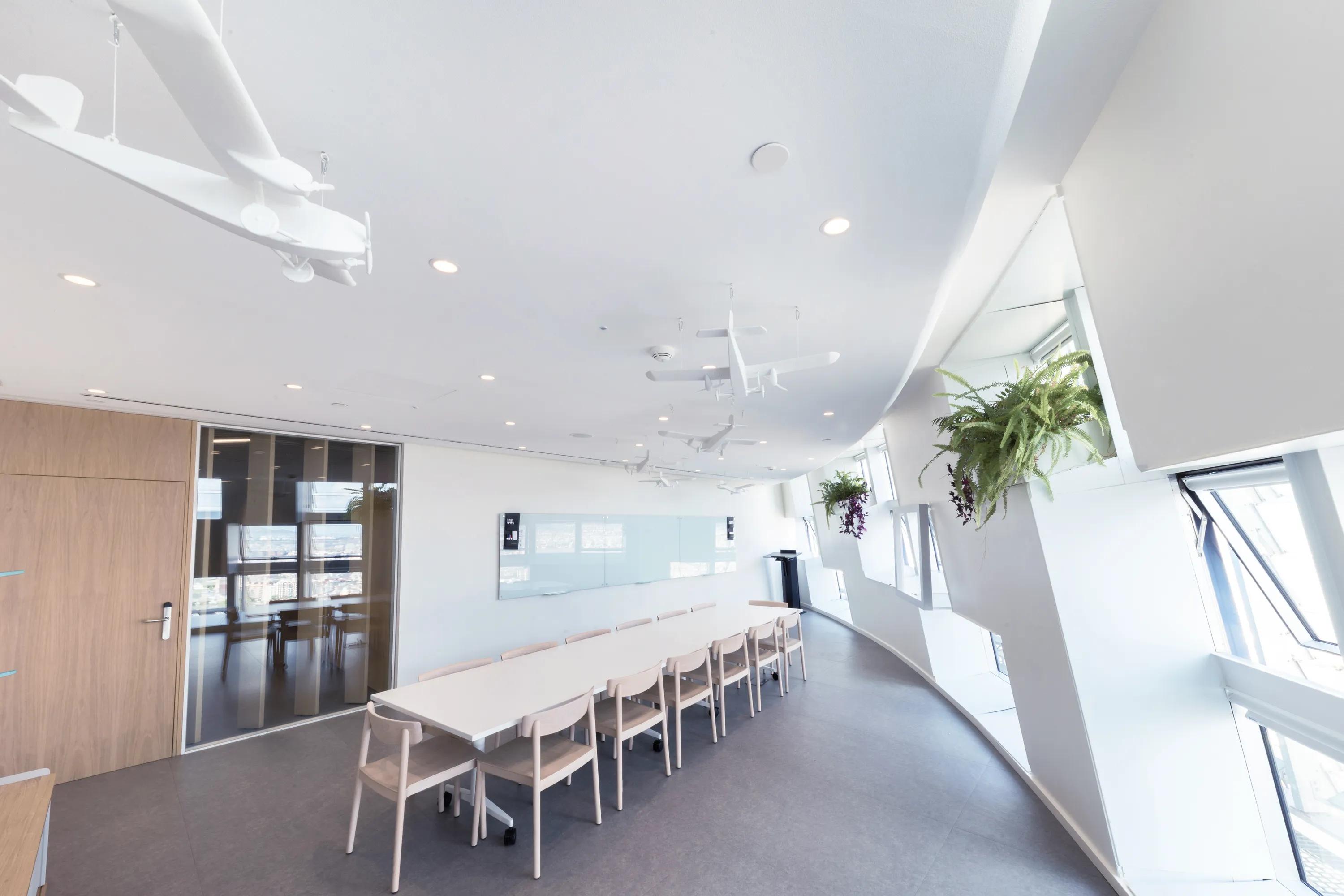 Meeting room with a curved monolithic acoustic ceiling, long table with chairs, suspended model planes, and angled windows bringing in natural daylight.