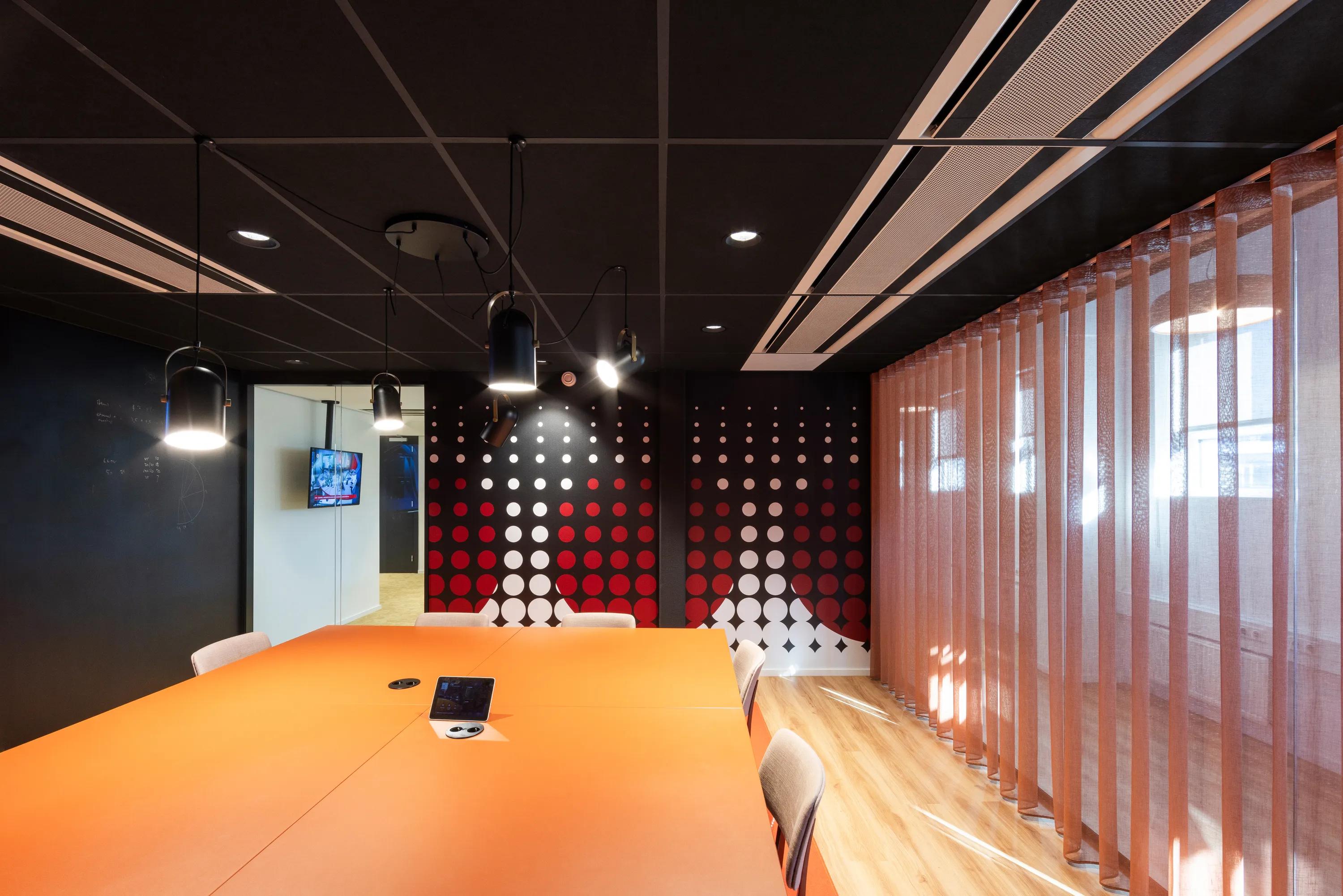 Meeting room with a dark acoustic grid ceiling, orange conference table, pendant lighting, and patterned feature wall, with sheer curtains filtering daylight along one side.