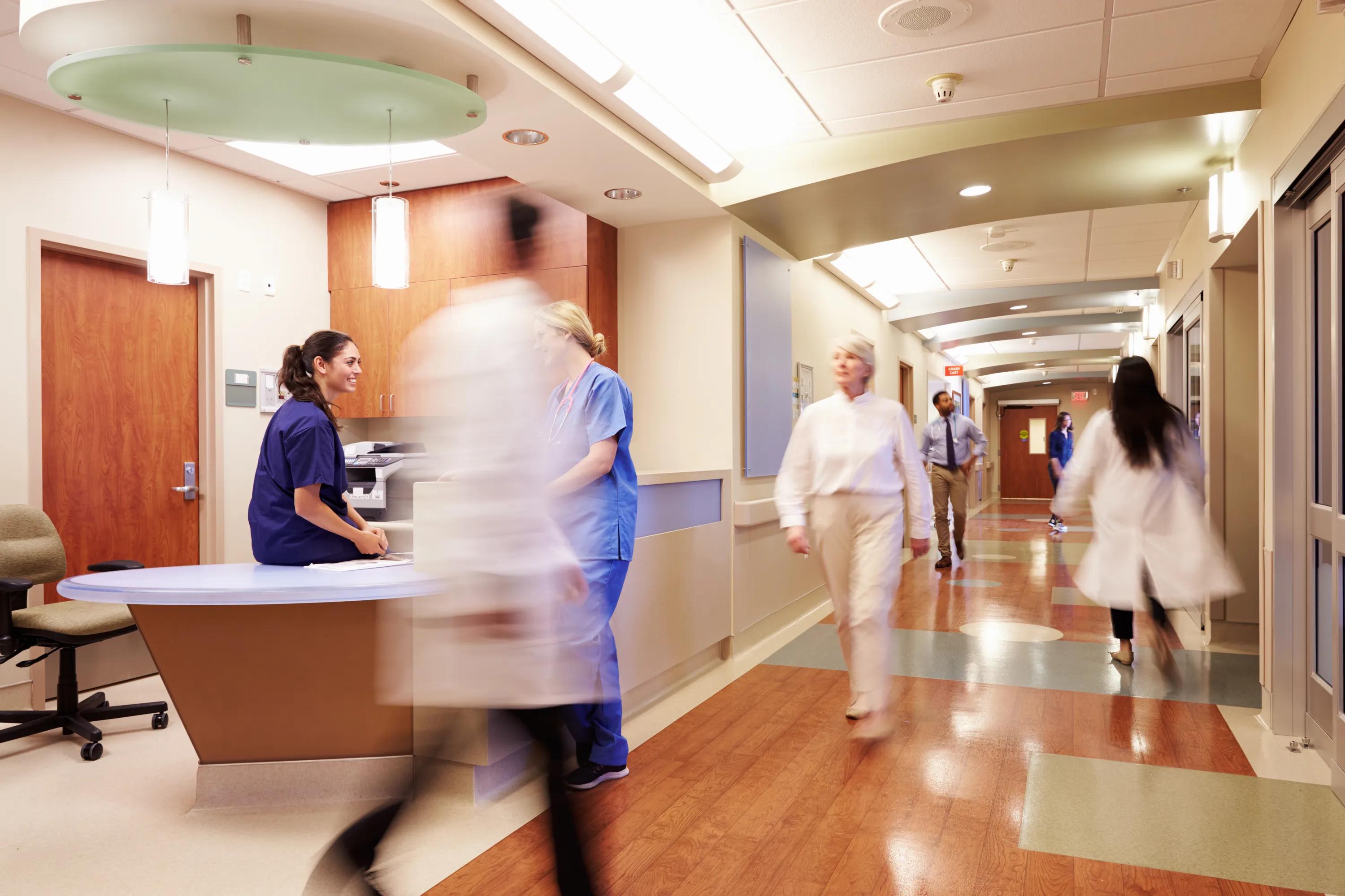Hospital corridor with nurses and doctors walking past a central reception desk, featuring a suspended circular acoustic panel above.