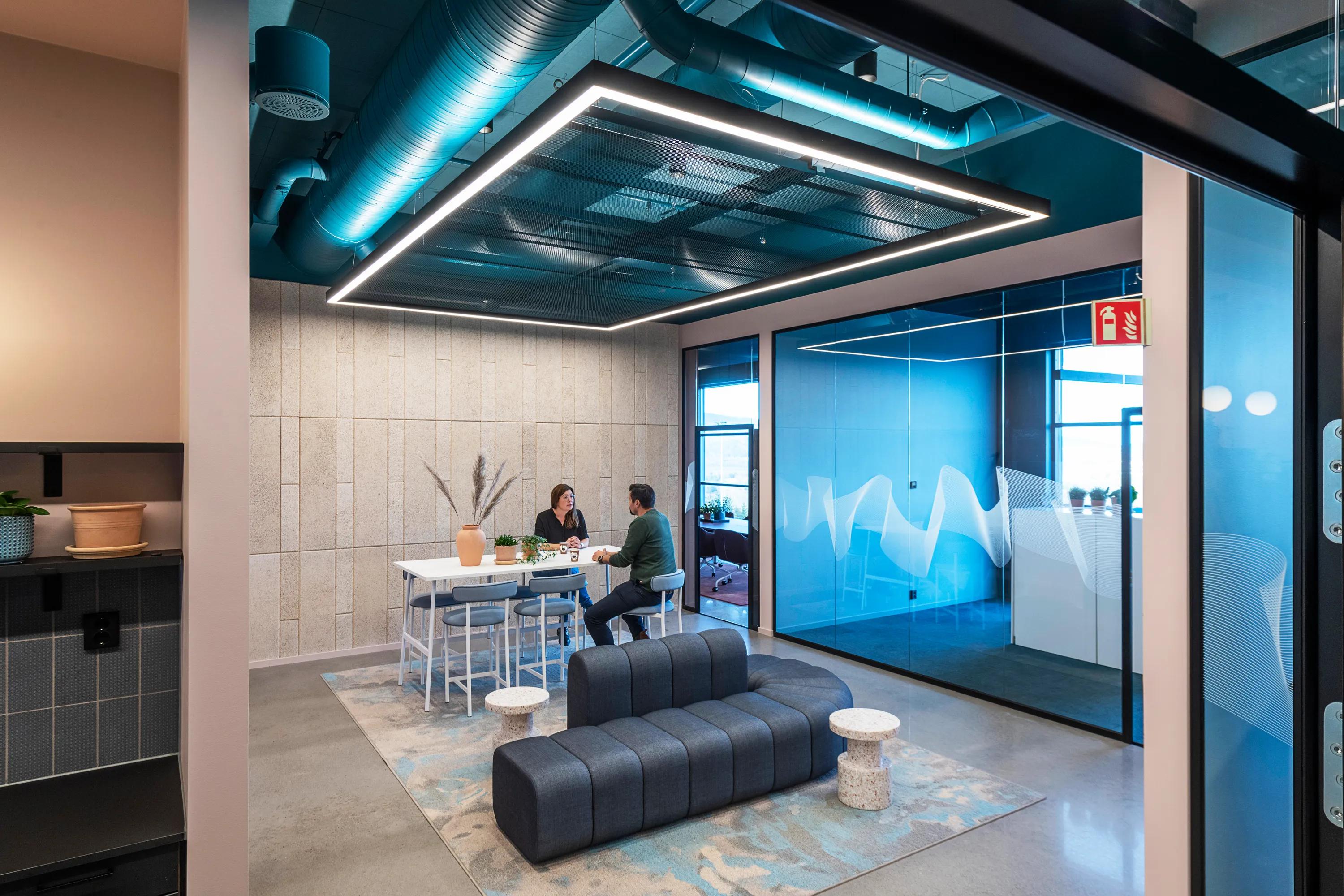 Modern office lounge with two people sitting at a high table, an exposed open plenum blue office ceiling, blue accents, a cushioned bench, and large windows.