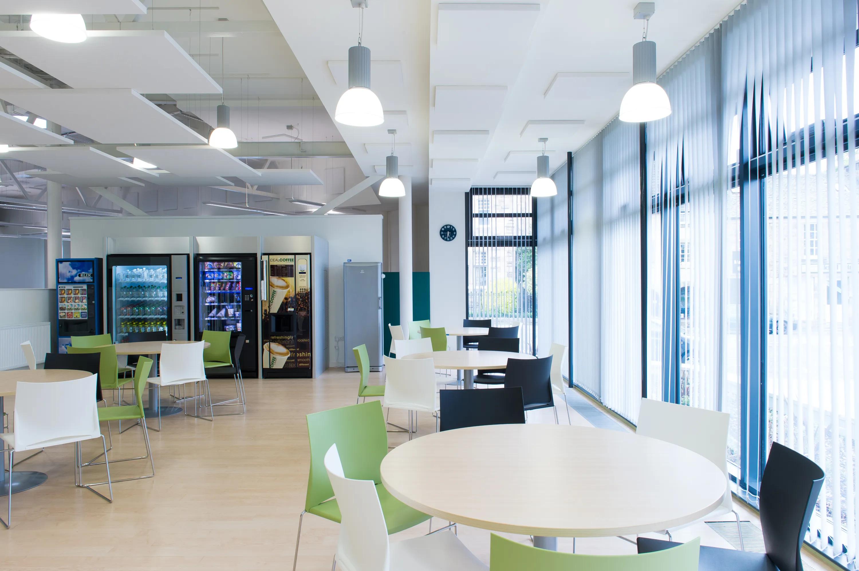 A modern, spacious break room with round tables, mixed chairs, vending machines, bright lighting, and large windows with vertical blinds.