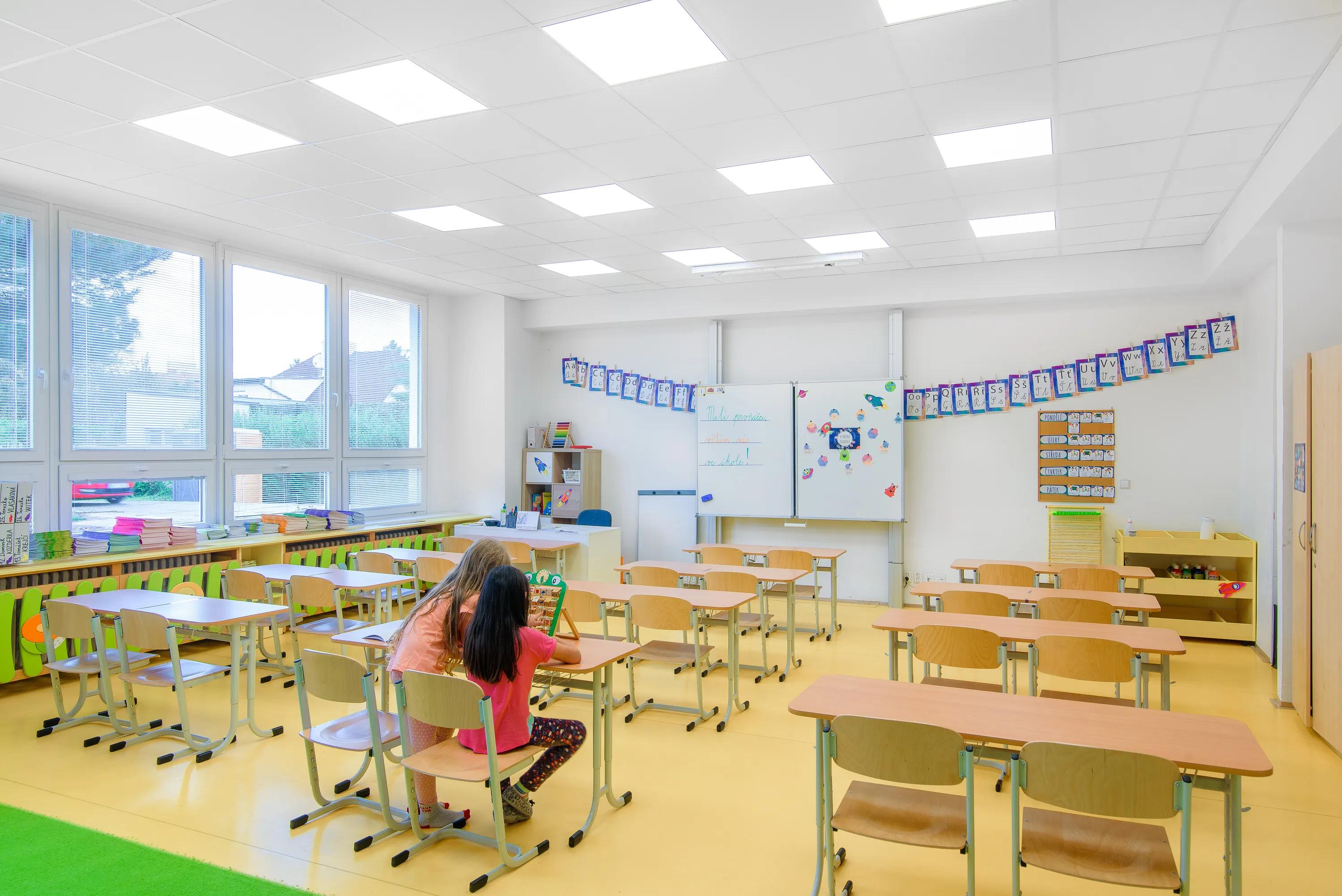 A colourful primary classroom featuring a white acoustic ceiling tile grid and bright yellow flooring, with two children working together at the front and large windows bringing in natural light.