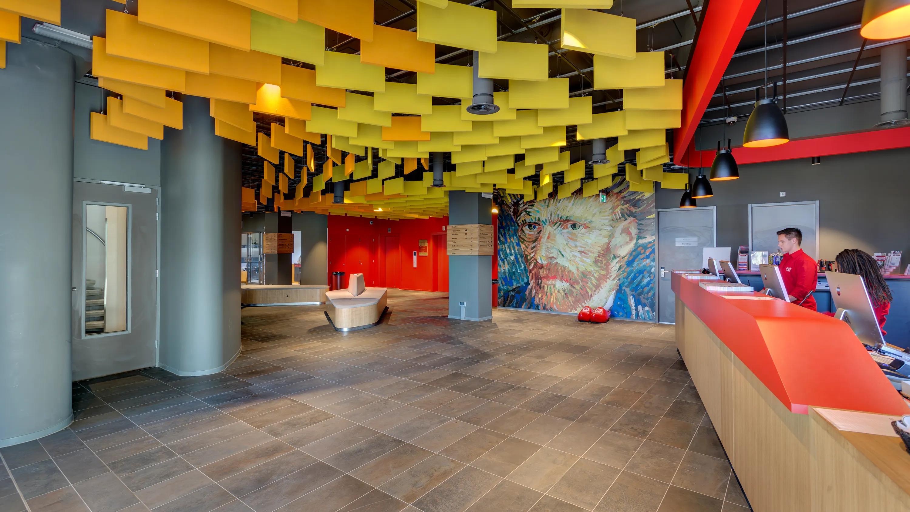 Reception area with suspended yellow acoustic baffles forming a dynamic ceiling, red reception desk, and large mural artwork on the wall behind the lobby space.