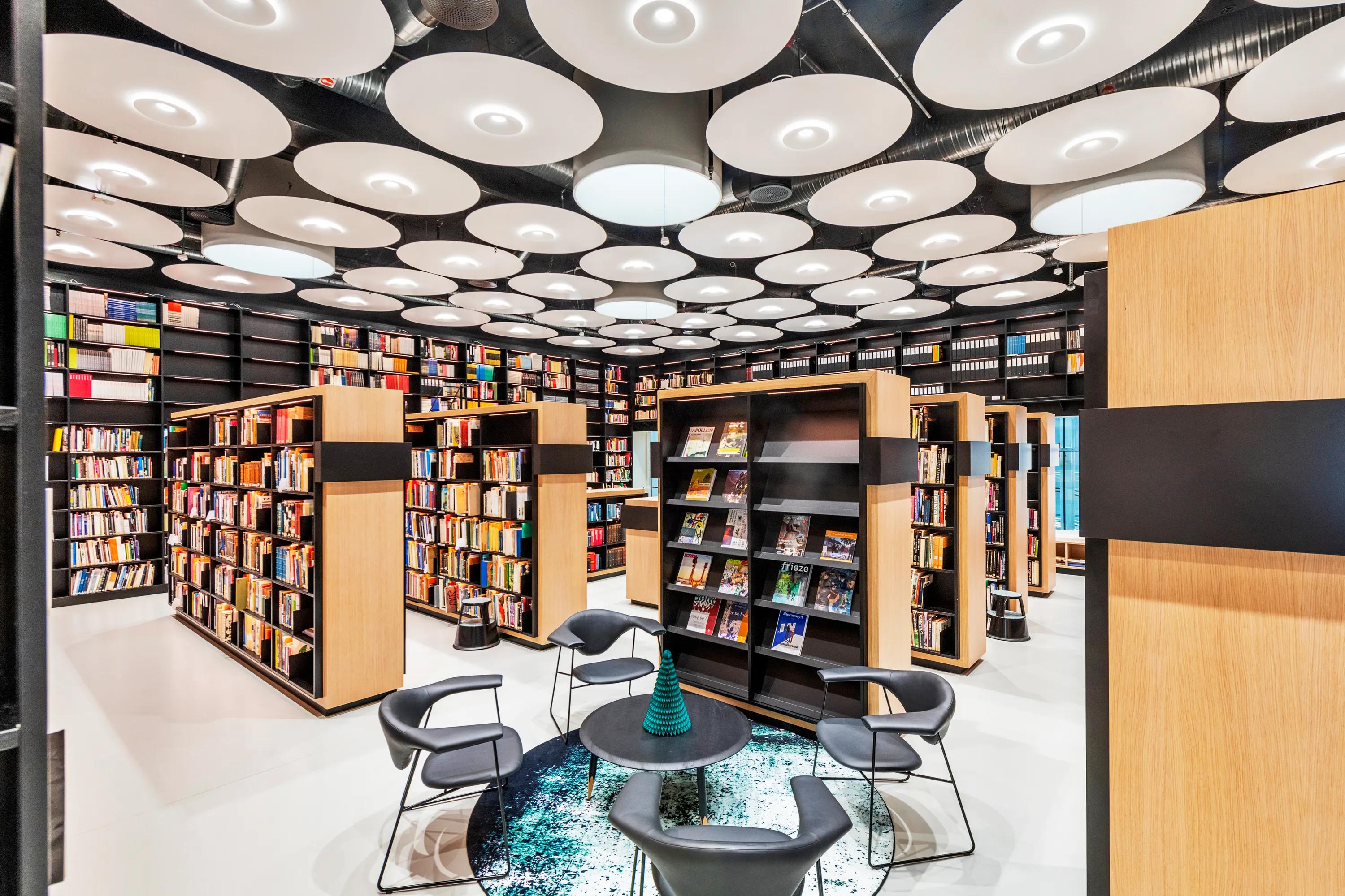A vibrant library interior featuring numerous circular white acoustic ceiling islands arranged across a dark soffit, with timber bookcases and lounge seating below.