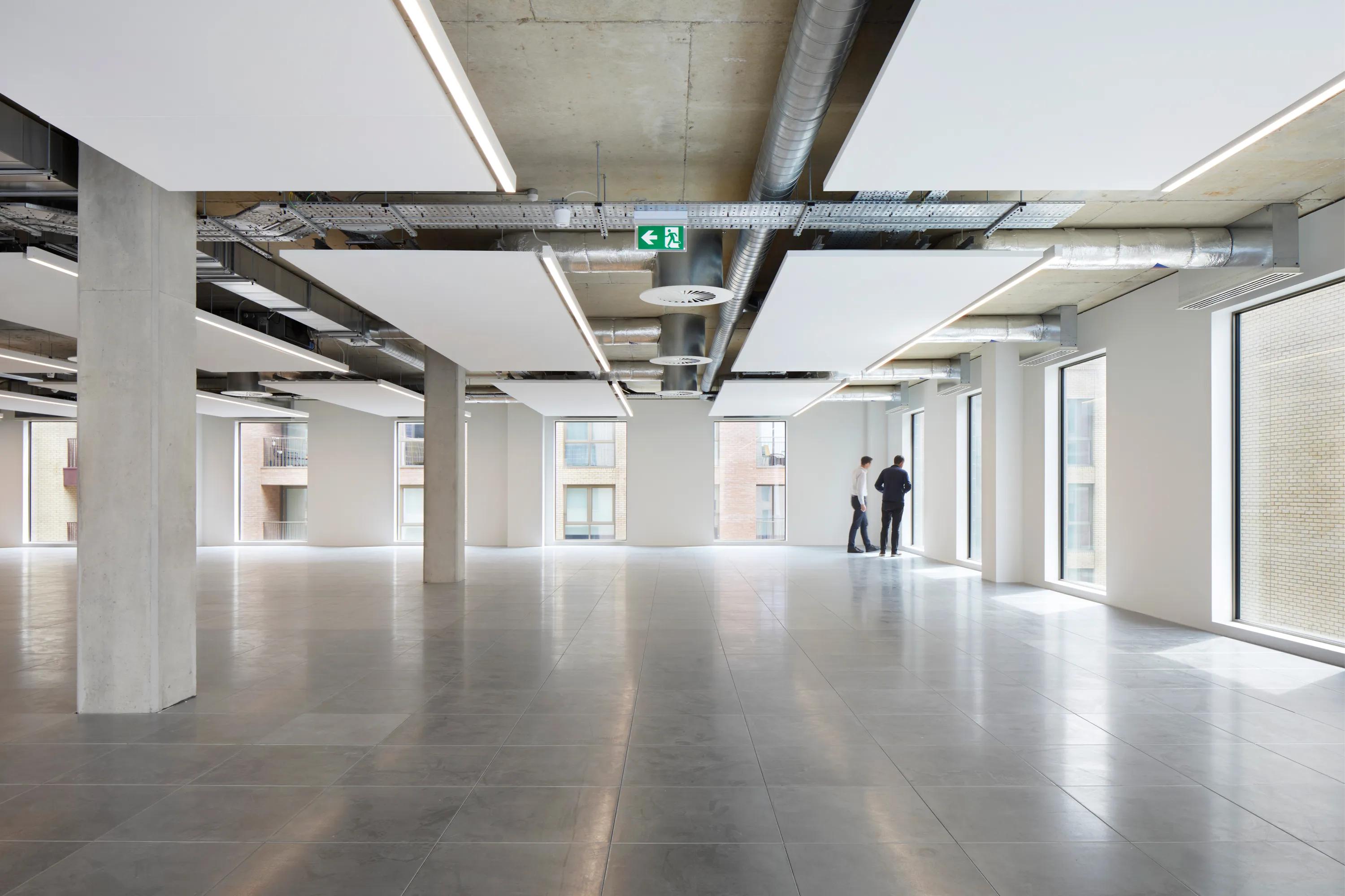 Spacious modern office with a floating raft ceiling, large windows, exposed ducts, and gray tile flooring. Two people stand near the window.