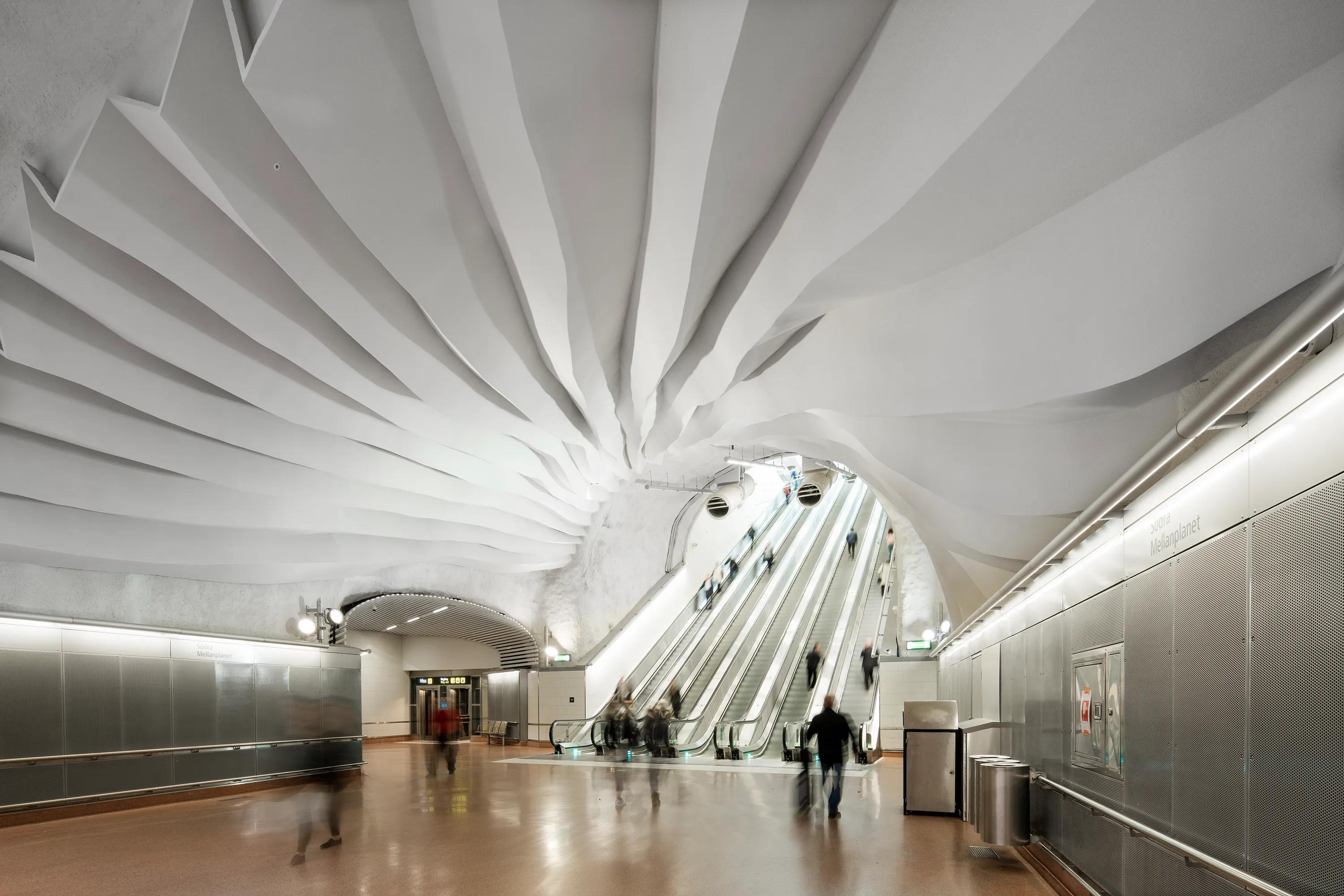 Stockholm metro station with a sculpted Rockfon Mono Acoustic ceiling forming a continuous artistic installation, escalators leading to the platform, and passengers moving through the space.