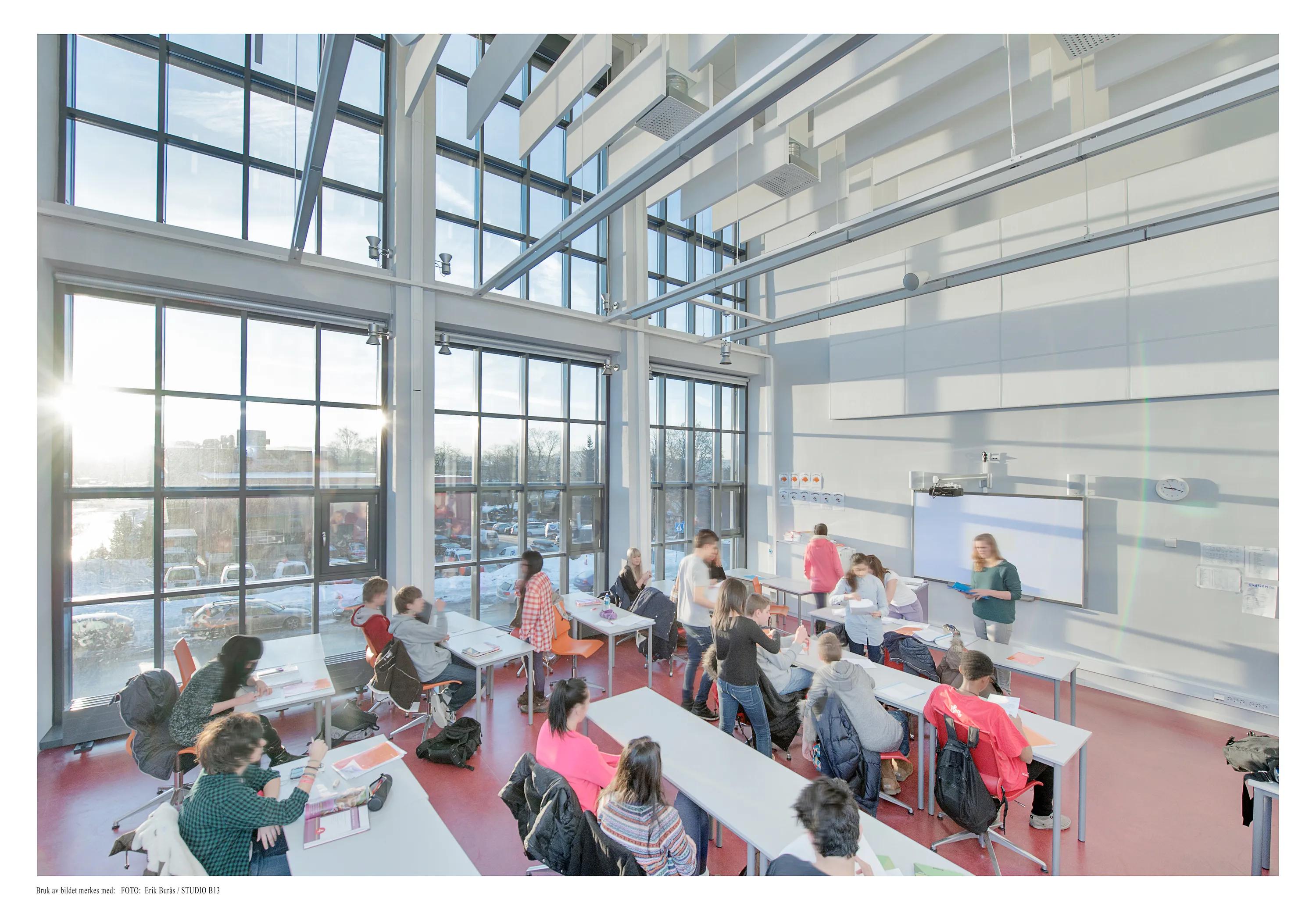 A large, light-filled classroom featuring suspended white acoustic baffles and exposed steel beams, with students working at tables below expansive floor-to-ceiling glazing.