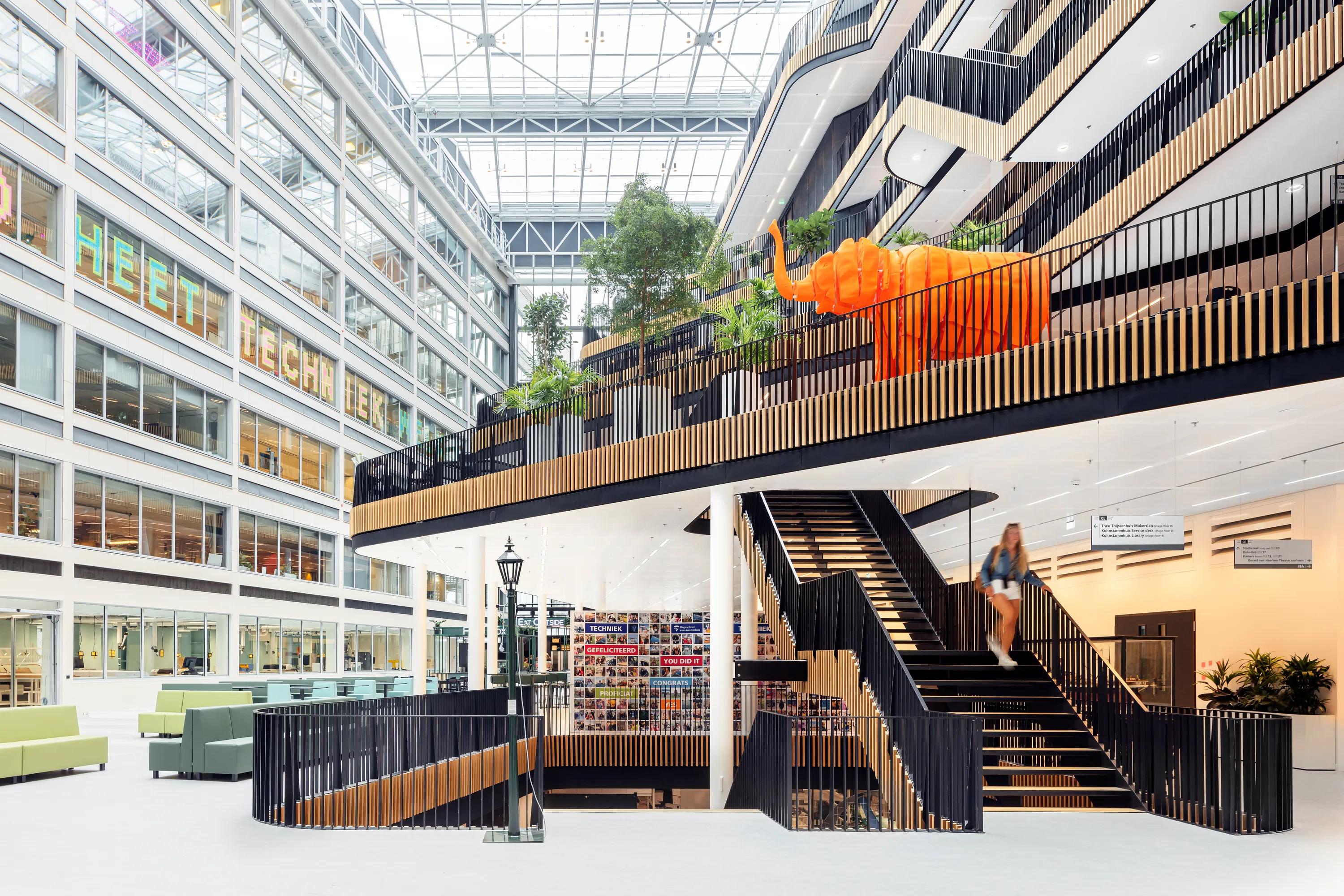 Large office atrium with a glass roof, central staircase, tiered walkways with planting, and an open, multi-level workspace filled with natural daylight.