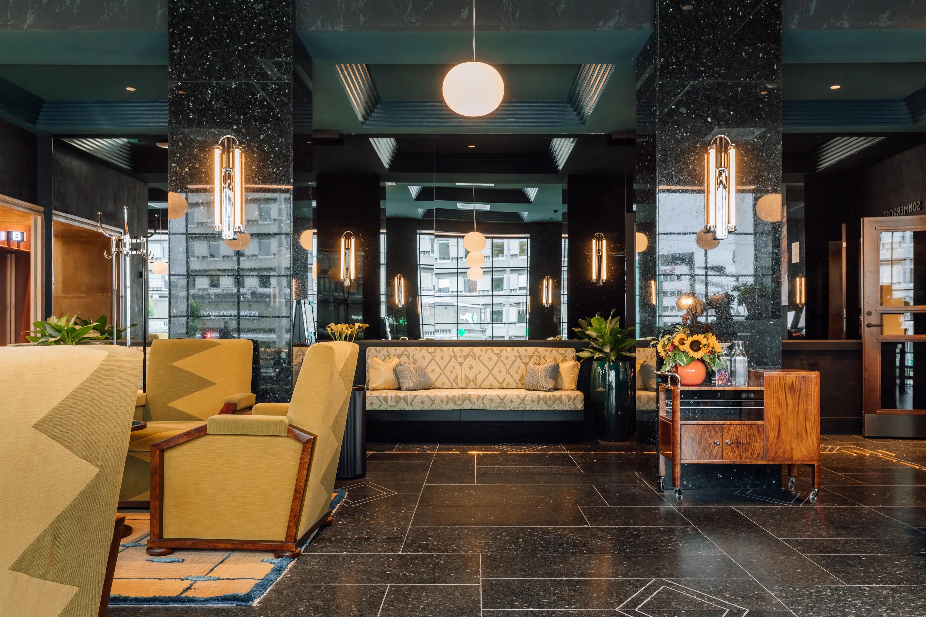 Hotel lounge with a coffered acoustic ceiling in dark blue, marble columns, seating areas, and decorative lighting creating a refined, symmetrical interior space.