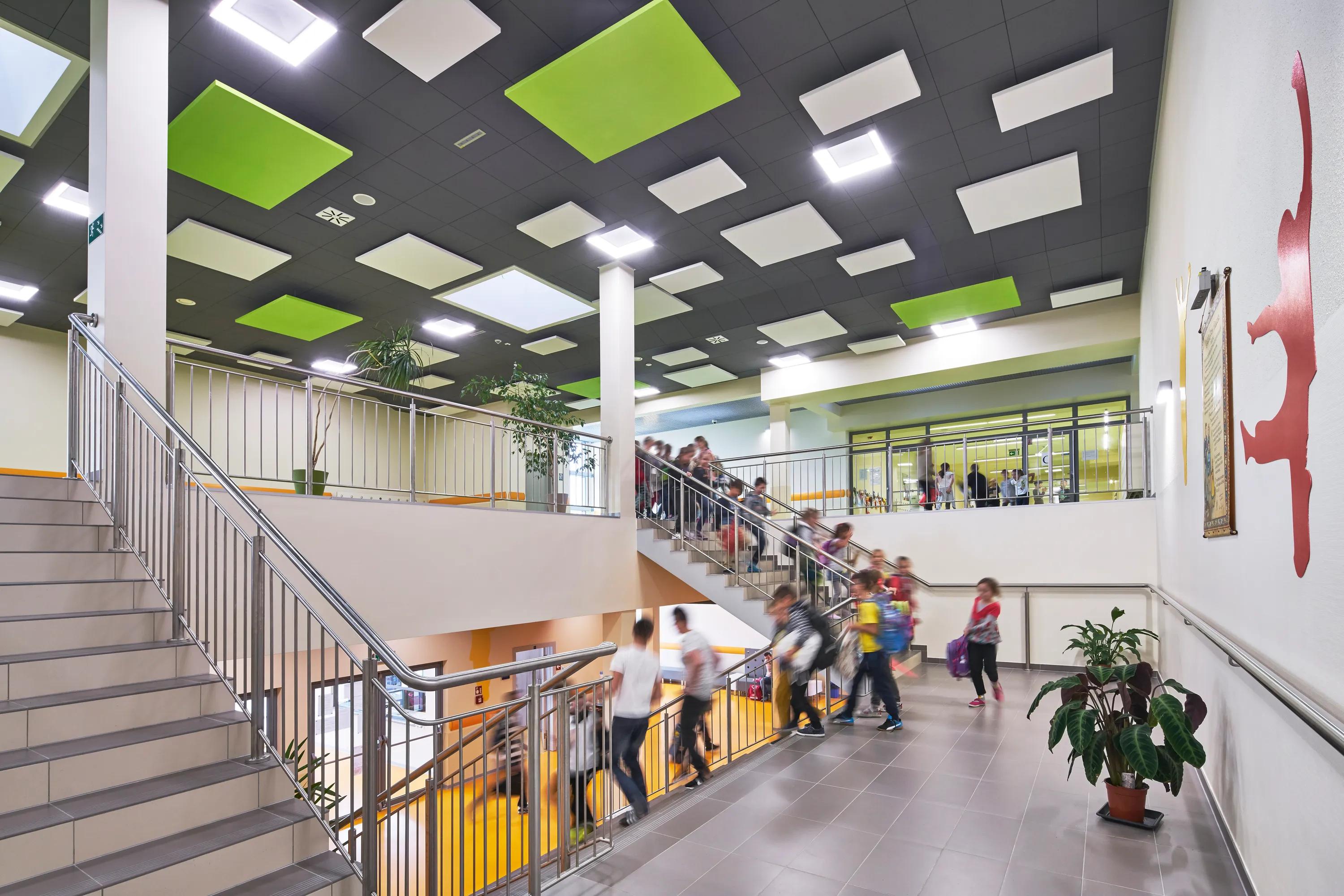 A modern school interior with students moving on a stairway. The ceiling features green and white panels, and there's a plant near the stairs.