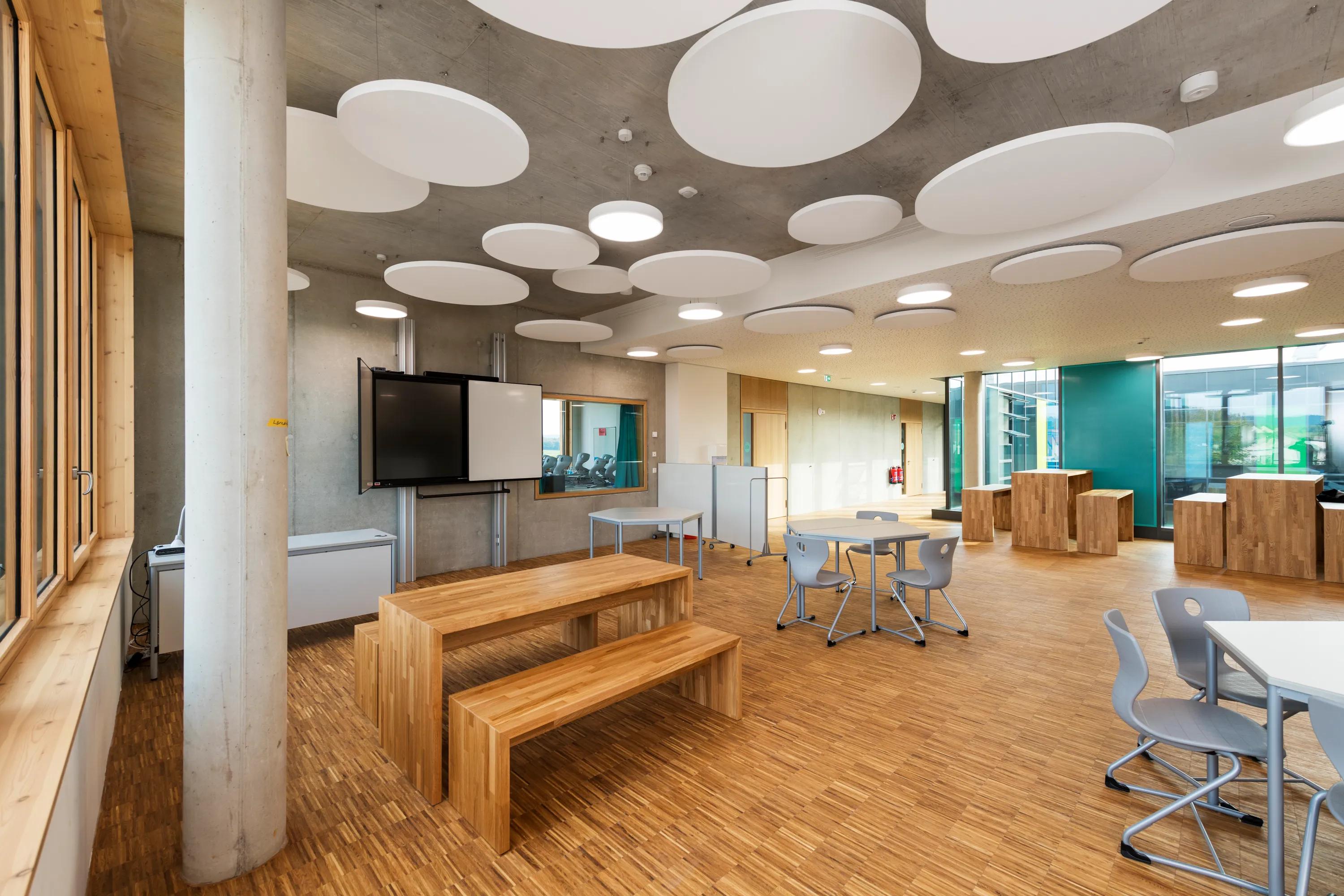 Modern classroom with wooden floors, benches, circular ceiling panels, a large screen, and blue-green walls. Natural light from large windows.