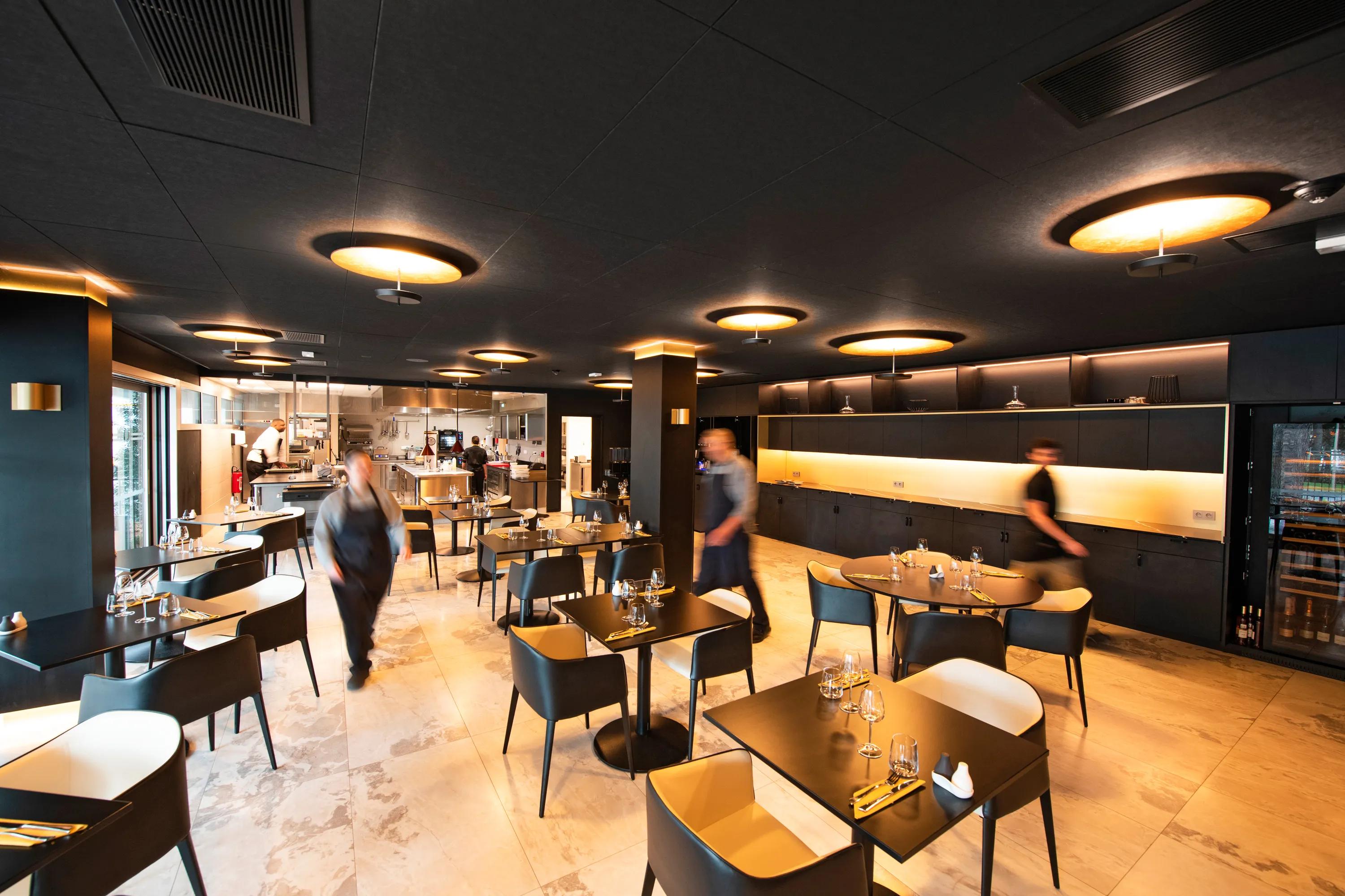 Modern restaurant interior with black suspended ceiling, several tables and chairs, dim lighting, warm tones, and blurred figures of staff walking.