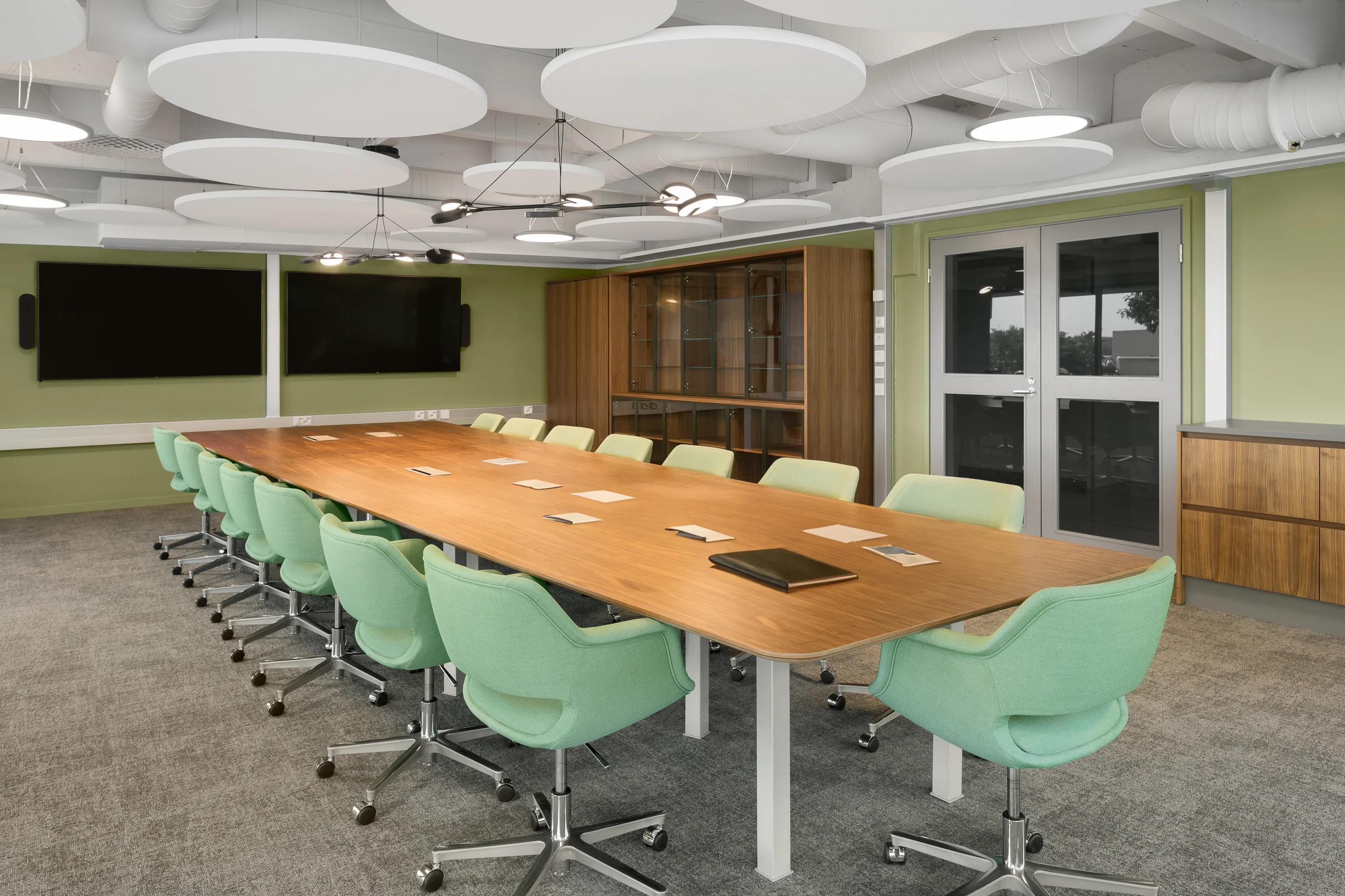 Meeting room with suspended circular acoustic rafts, large conference table, green chairs, and integrated screens in a modern office setting.