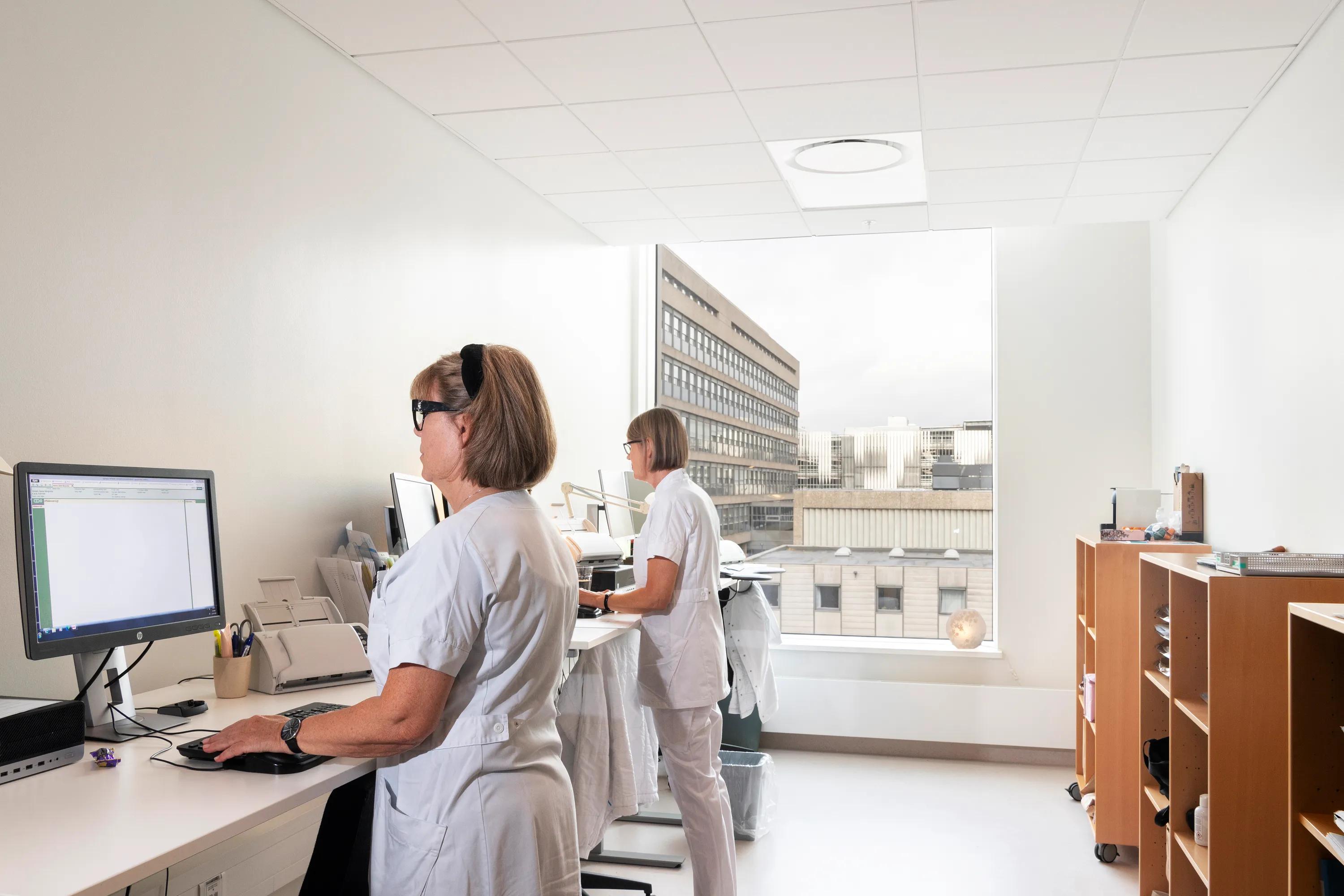 Two healthcare staff working at computers in a bright clinical office with a white acoustic ceiling and large window overlooking adjacent hospital buildings.