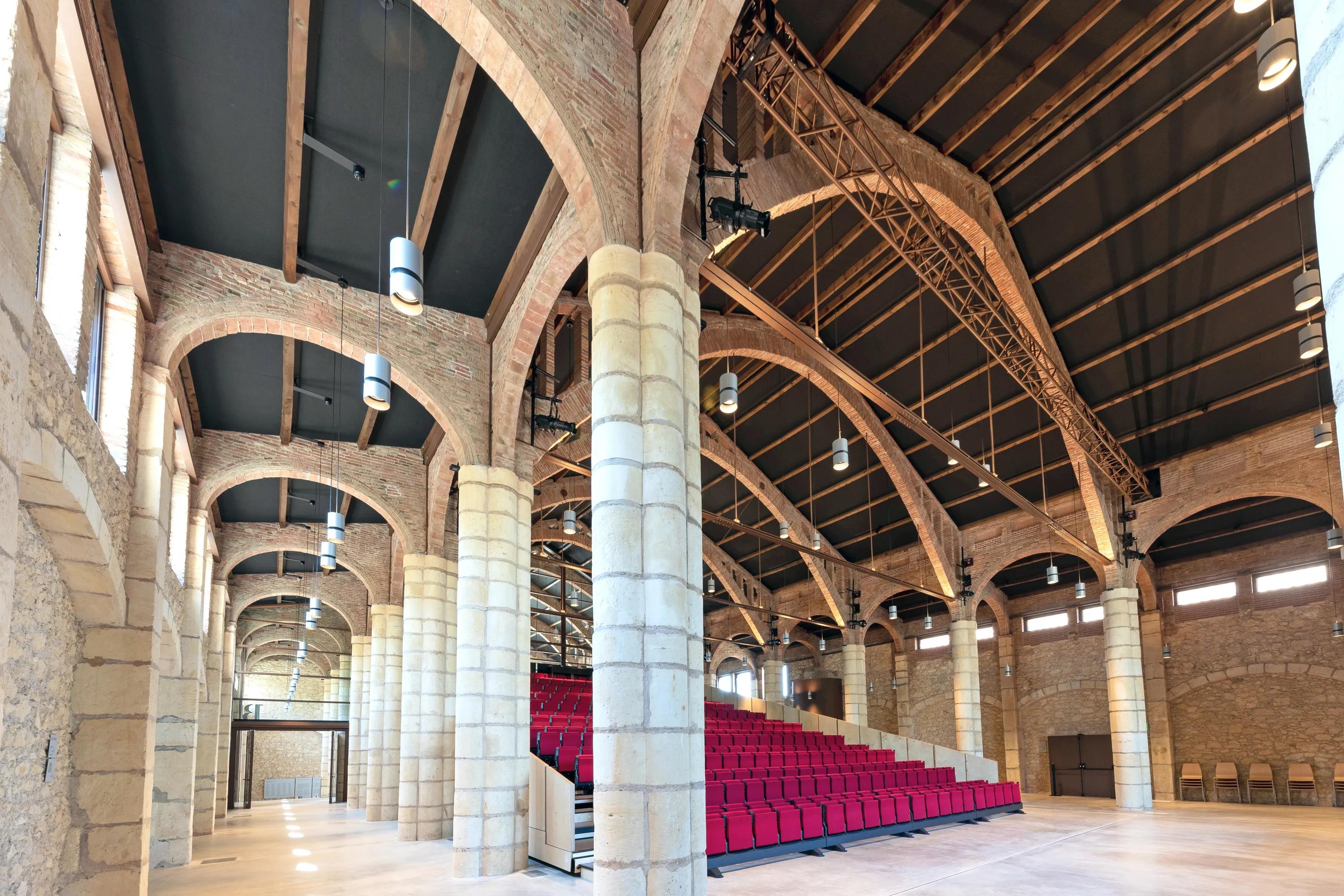 Auditorium with tiered red seating, a stage area, exposed brick arches, and black acoustic ceiling tiles fitted between the vaulted roof beams.