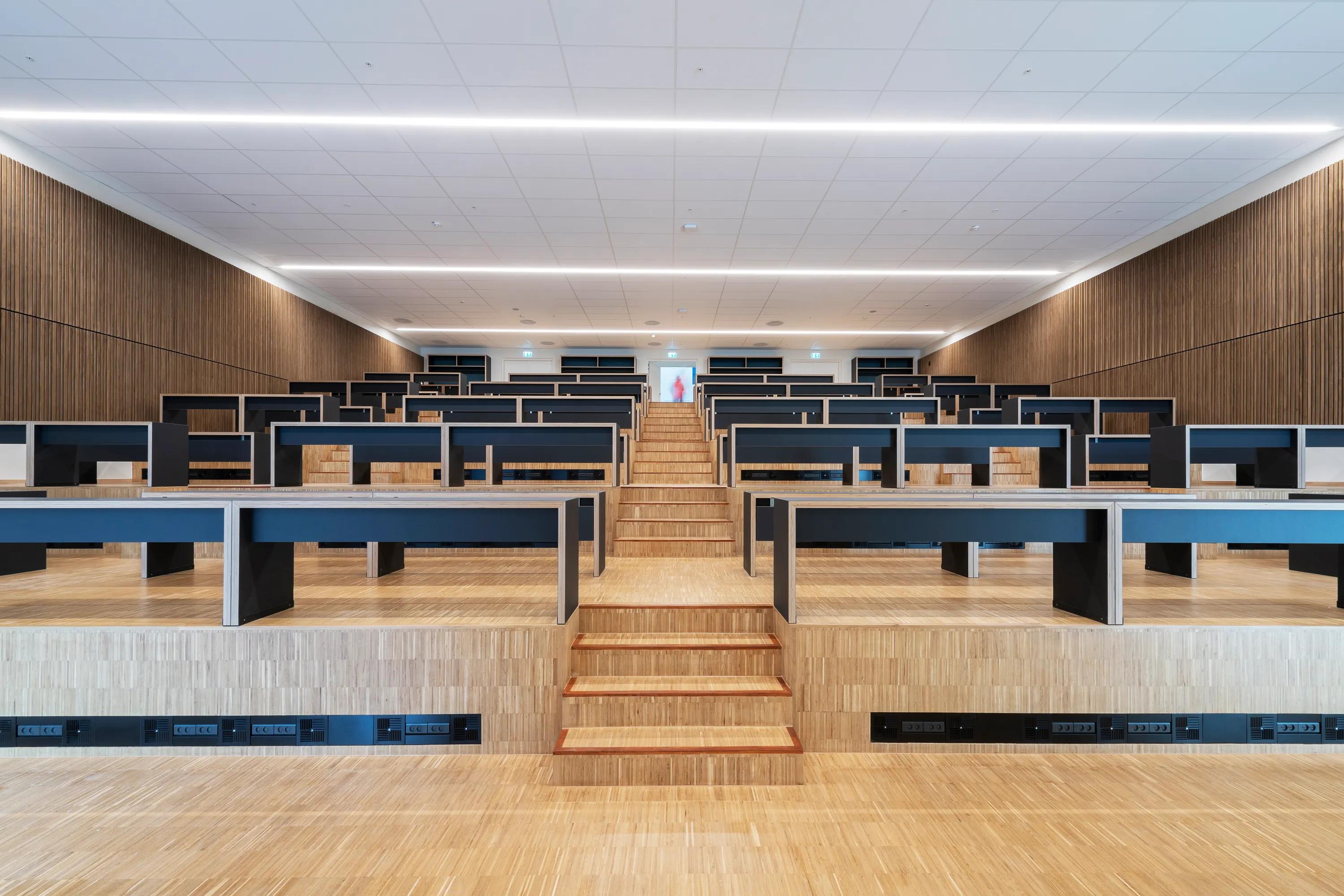 A spacious tiered lecture hall featuring a white acoustic ceiling tile grid, stepped timber flooring, black study benches, and integrated lighting