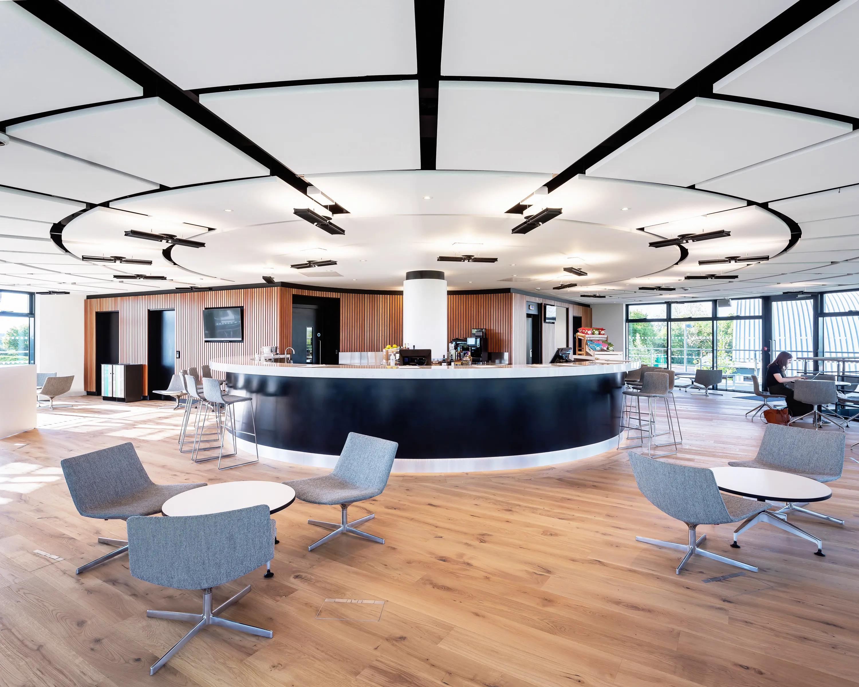 Modern open-concept lounge with floating raft ceilings, central circular counter, surrounded by gray chairs and small tables on a wooden floor, featuring large windows.