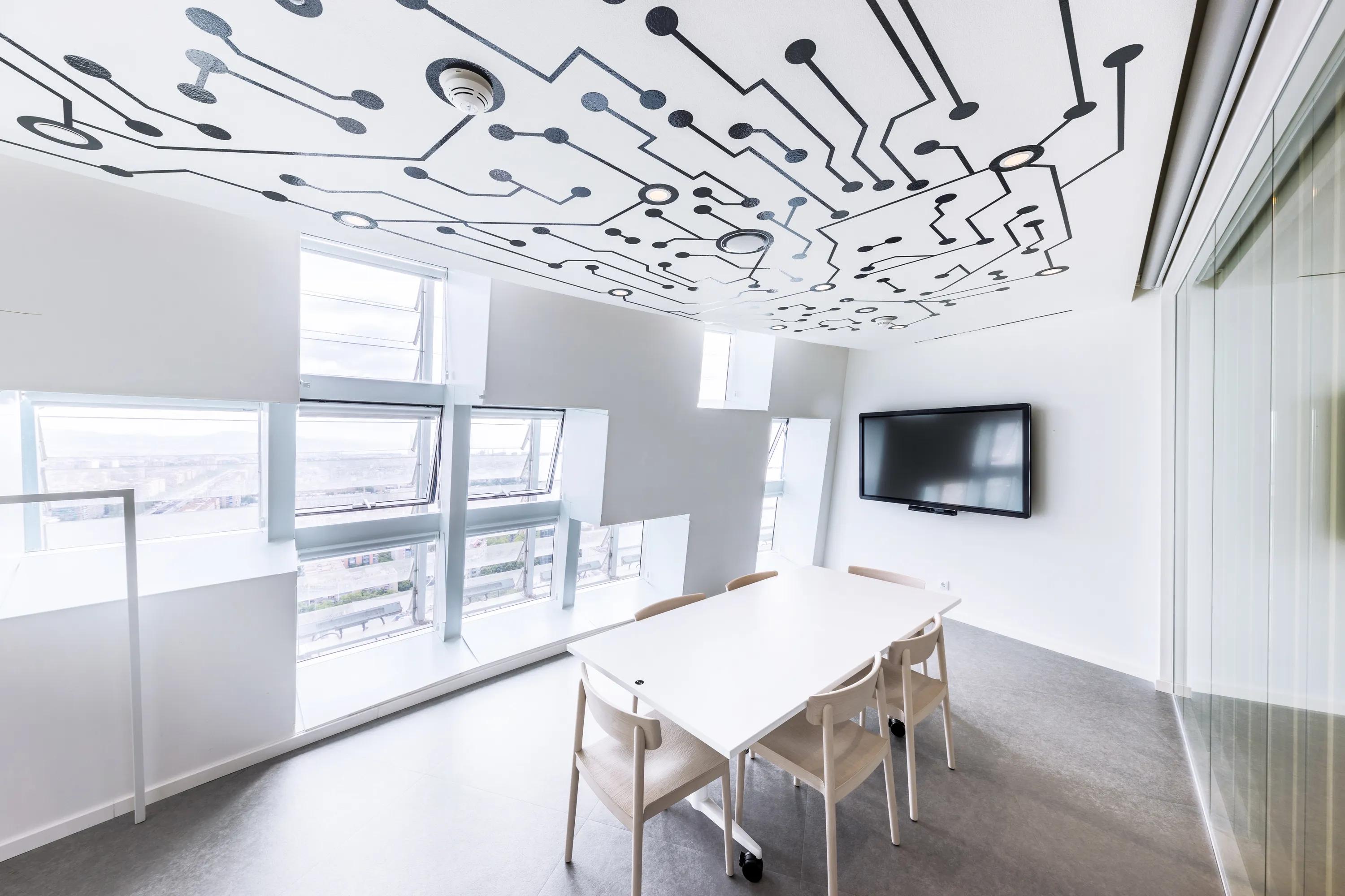Meeting room with acoustic ceiling tiles featuring a printed circuit pattern, table and chairs, wall-mounted screen, and angled windows bringing in natural daylight.