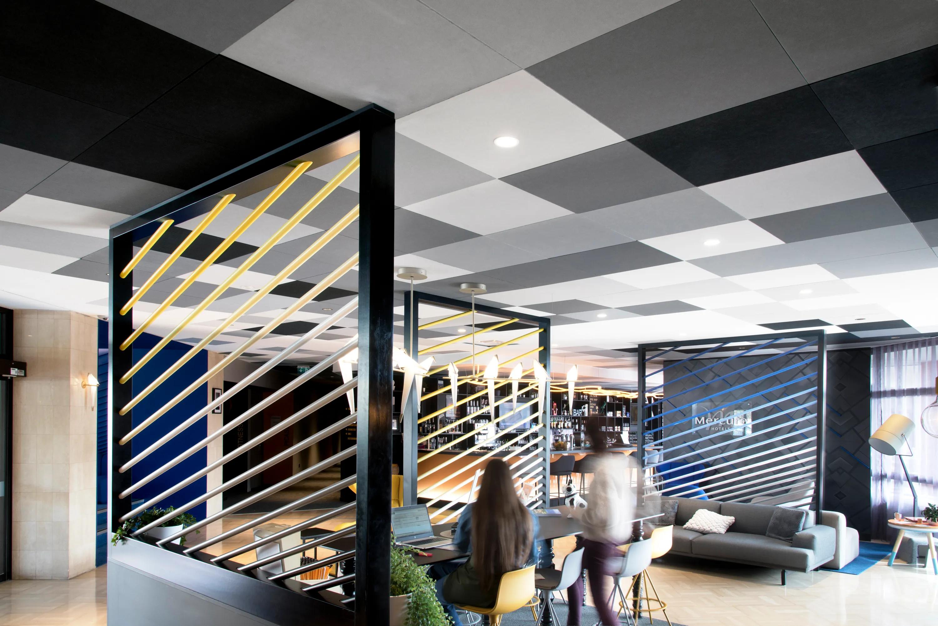 Hotel bar and lounge with a black and white checkerboard acoustic ceiling, illuminated bar counter, seating areas, and large windows bringing daylight into the space.