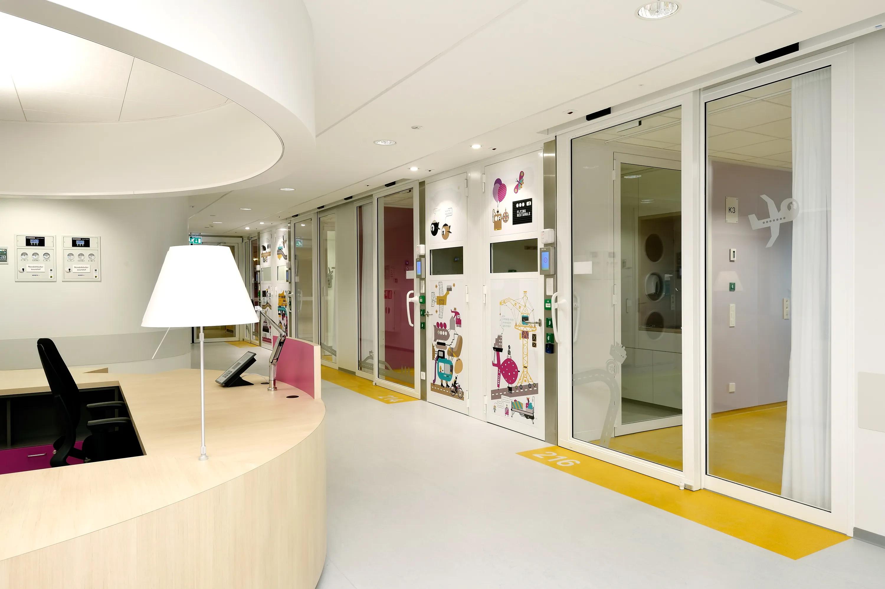 Children’s hospital corridor with a curved wooden nurse desk, bright white acoustic ceiling panels, and glass room doors decorated with colourful illustrations and icons.