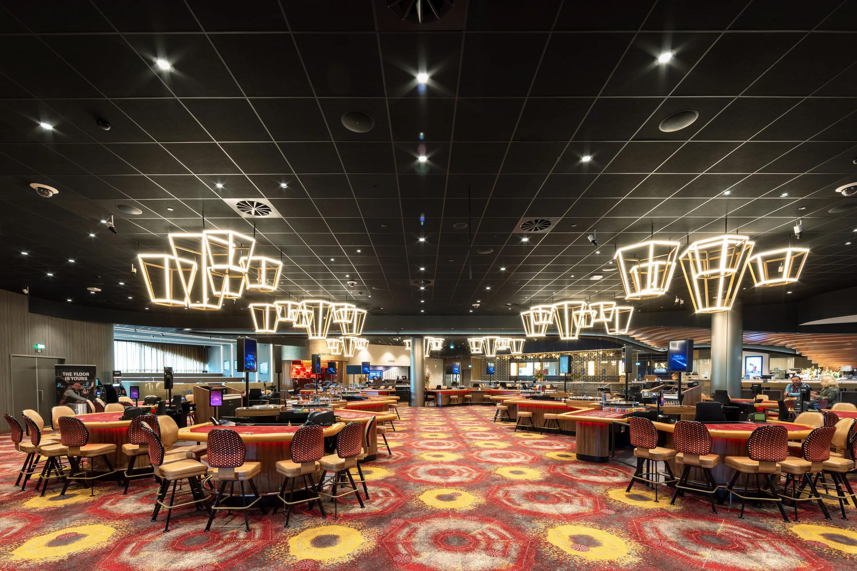 Spacious casino interior with a black suspended ceiling, modern geometric chandeliers, colorful patterned carpet, and rows of gaming tables and chairs.